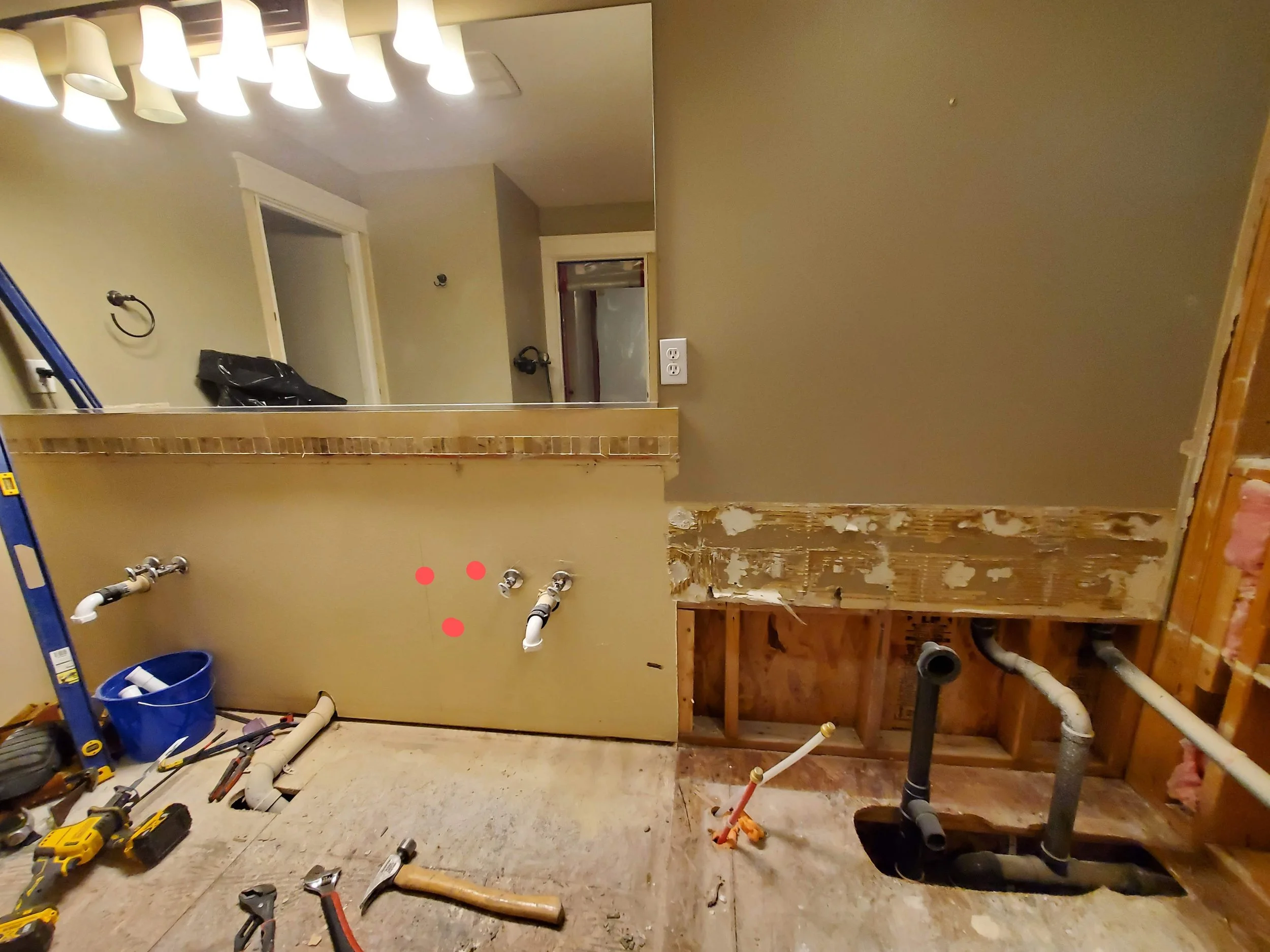 A bathroom under renovation with exposed pipes and partially installed wall panels, tools on the floor, and a large mirror reflecting ceiling lights.