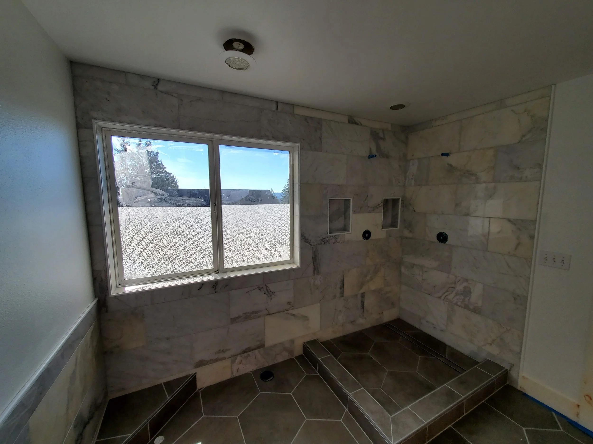 Bare bathroom with marble walls and dark tile flooring, window with frosted lower pane, and shower area with stone step and wall niches.