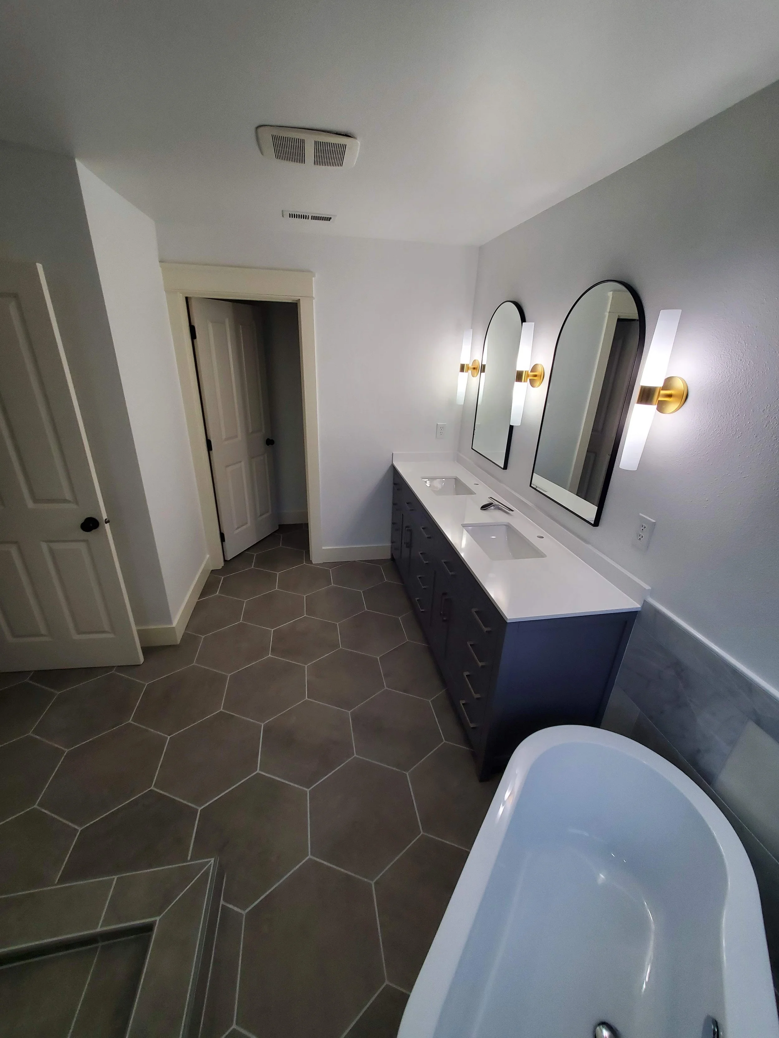 Modern bathroom with white walls, hexagon gray floor tiles, a navy blue vanity with two sinks, two arched mirrors, and two vertical wall sconces with white shades.