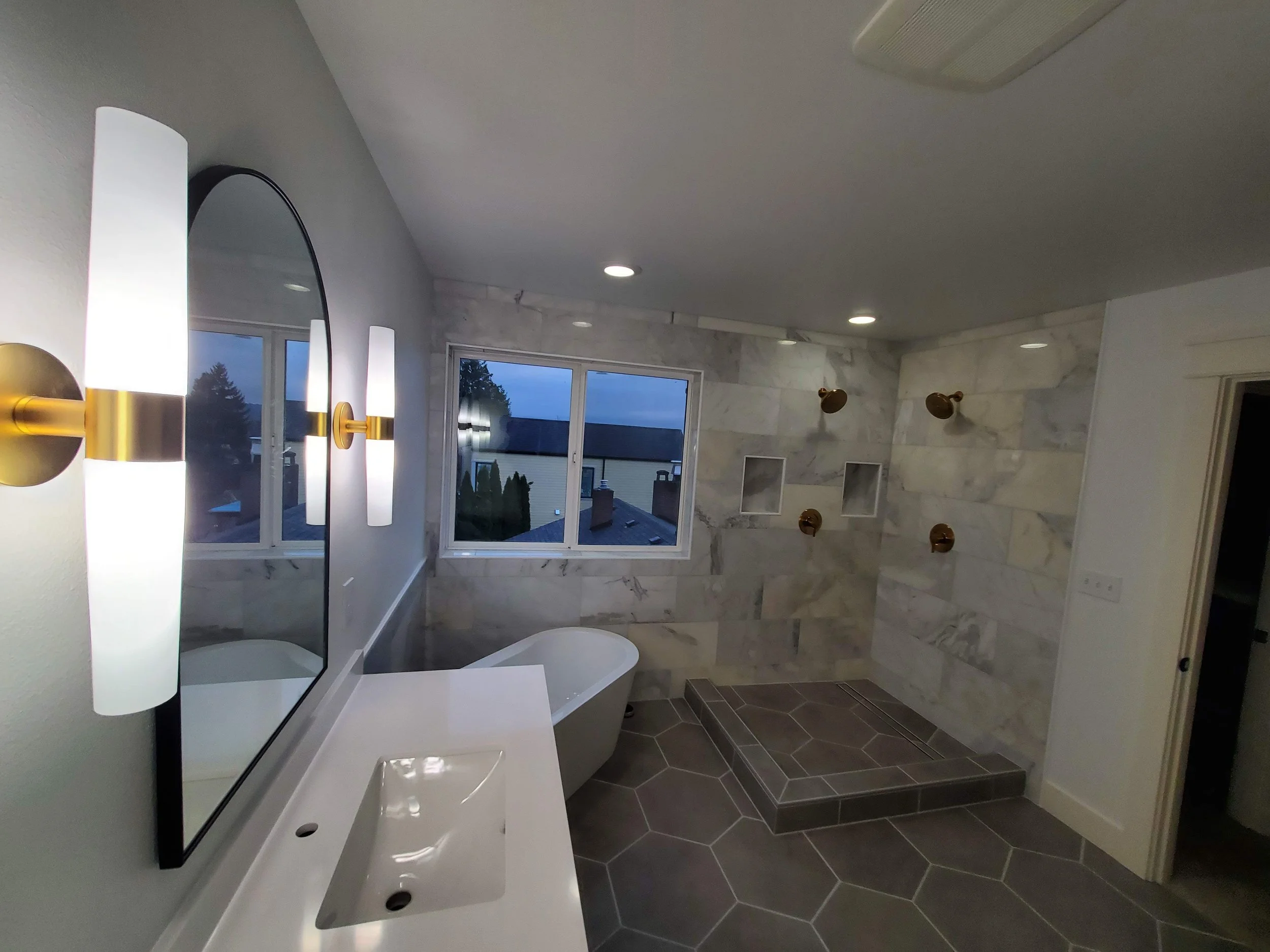Modern bathroom with marble walls, window view, wall-mounted light fixtures, a bathtub, a large mirror, and a walk-in shower with multiple showerheads.