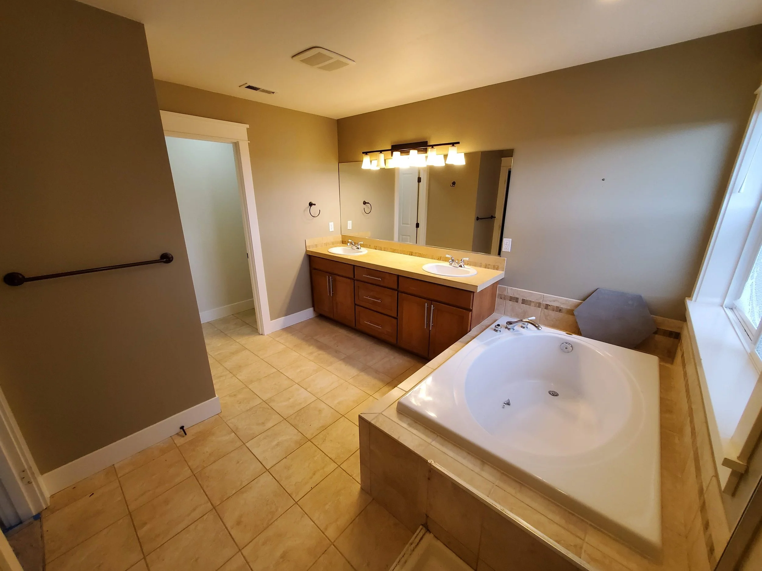 Bathroom with double vanity sinks, large mirror with light fixtures, corner bathtub with a window, beige tiled floor, and neutral wall color.