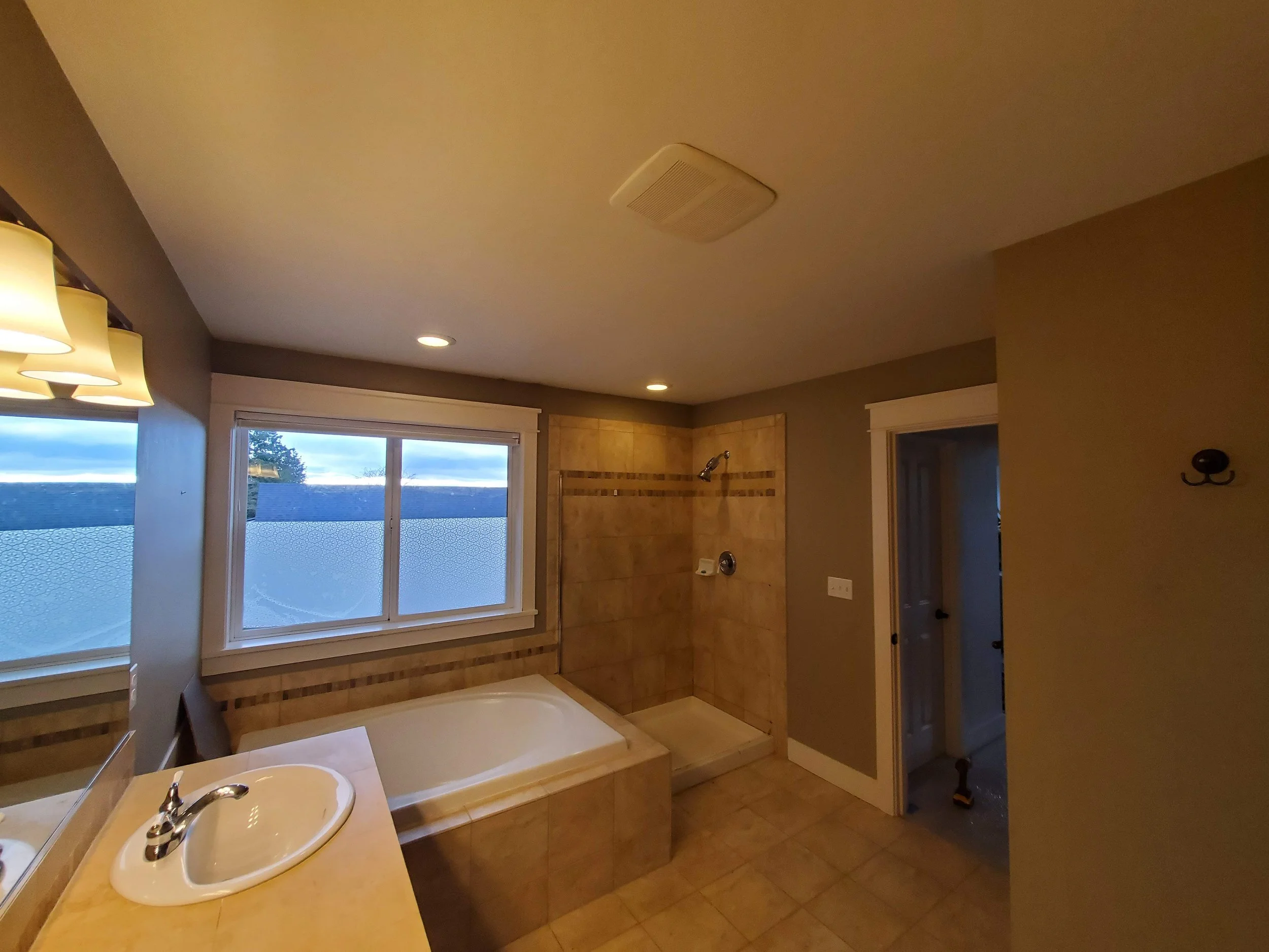 A bathroom featuring a window, a bathtub, a separate shower area, a small sink, and beige tiles, with brown walls and white trim.