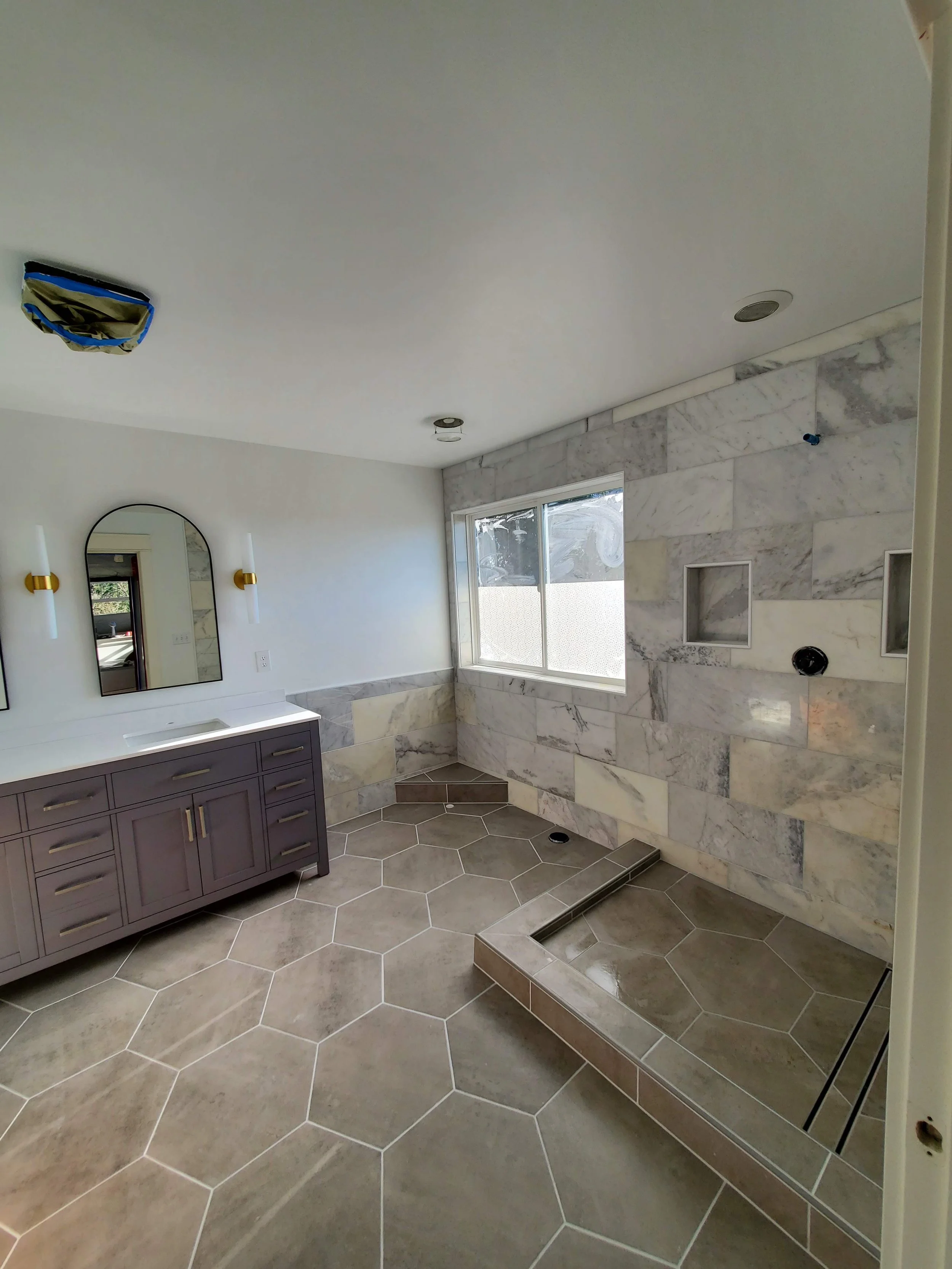 Bathroom under construction with hexagonal tile flooring, purple vanity with gold handles, arched mirror, wall-mounted lights, and a tiled shower area with a window and built-in niches.