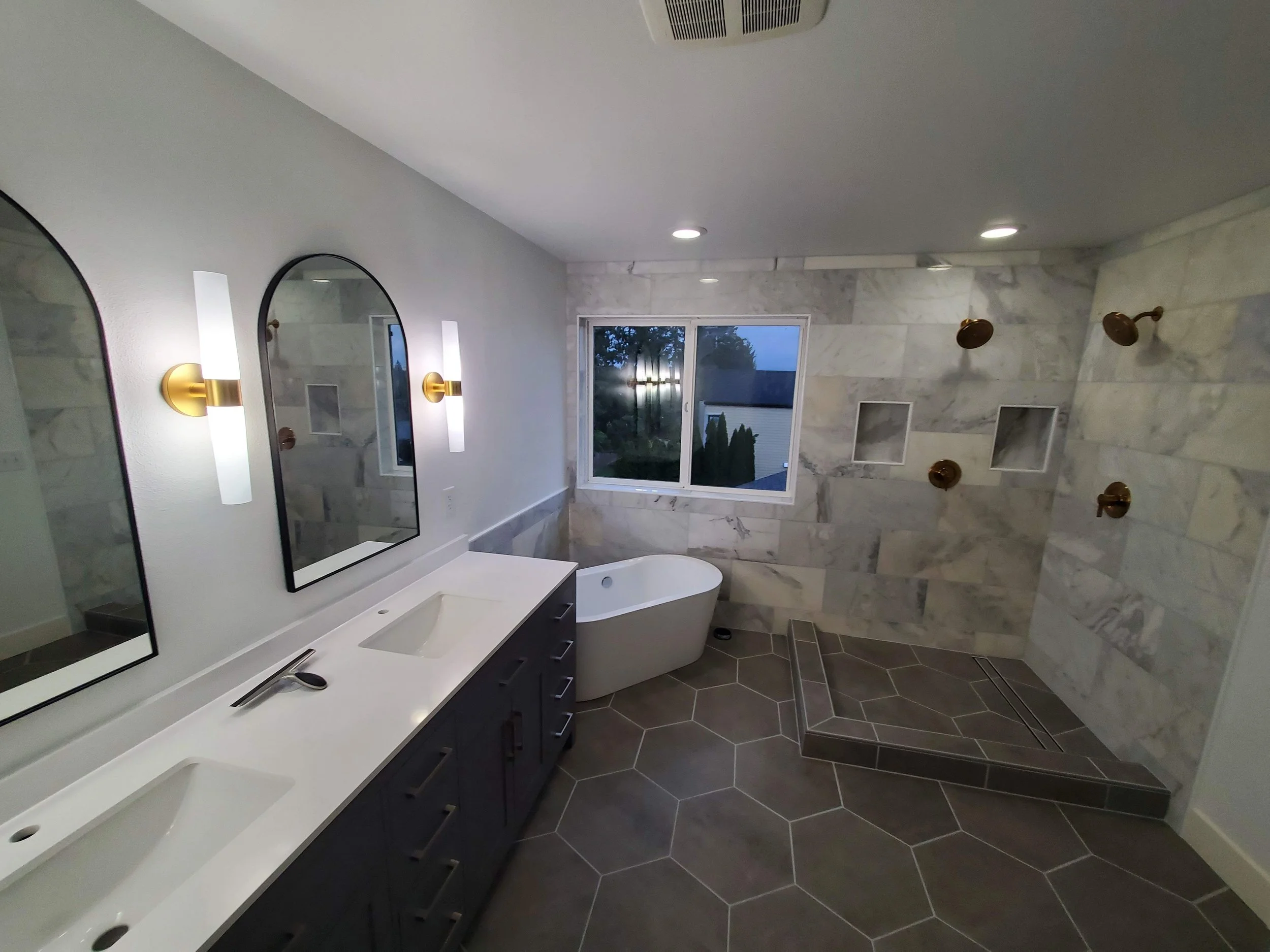 Modern bathroom with dual sinks, oval mirror, gold wall sconces, standalone bathtub, large window, and walk-in shower with multiple showerheads and shelves.