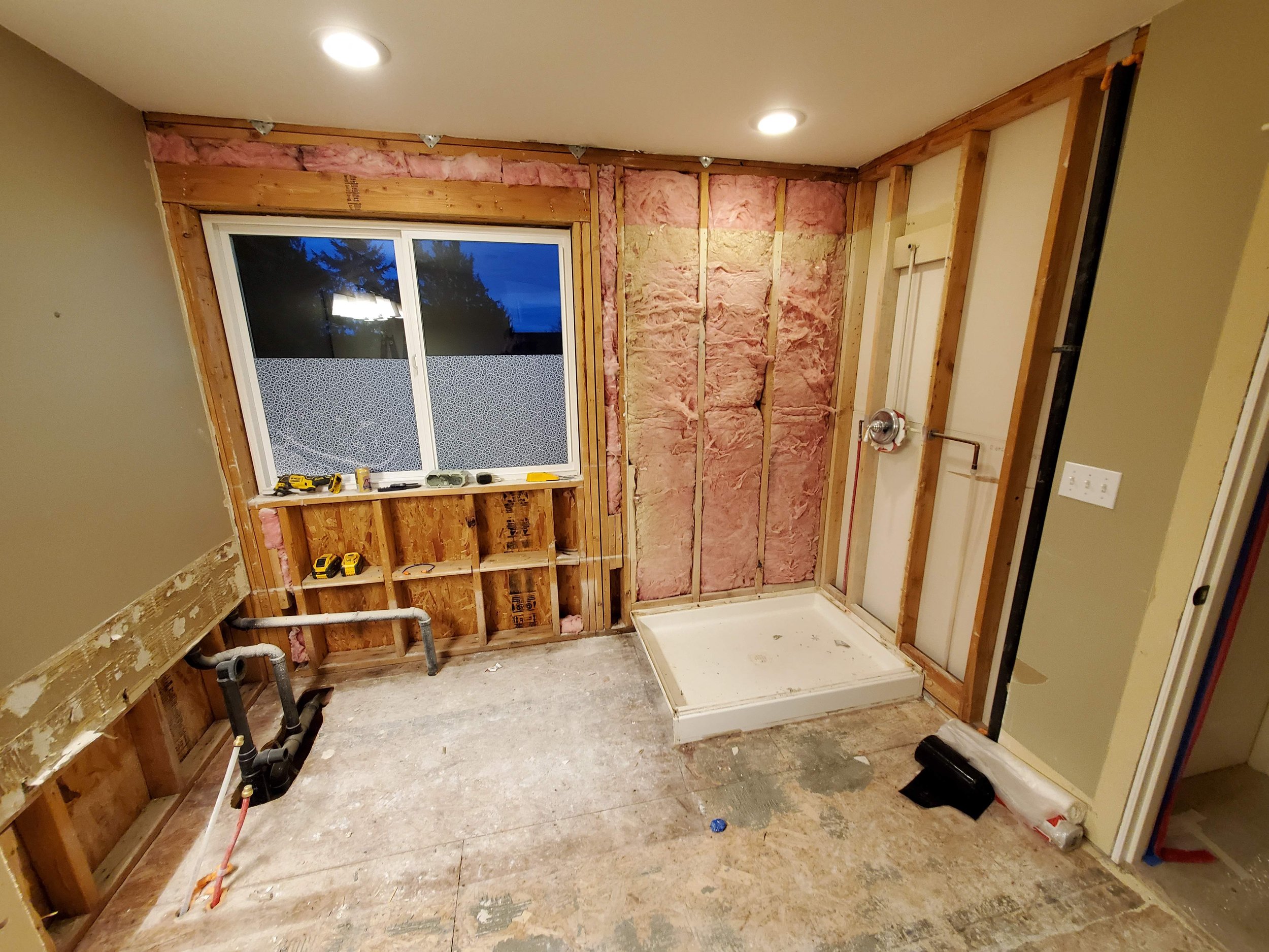 Bathroom under renovation with exposed wall studs and insulation, a window with tools on the window sill, and a shower base installed in the corner. Interior has unfinished walls, flooring, and visible plumbing.