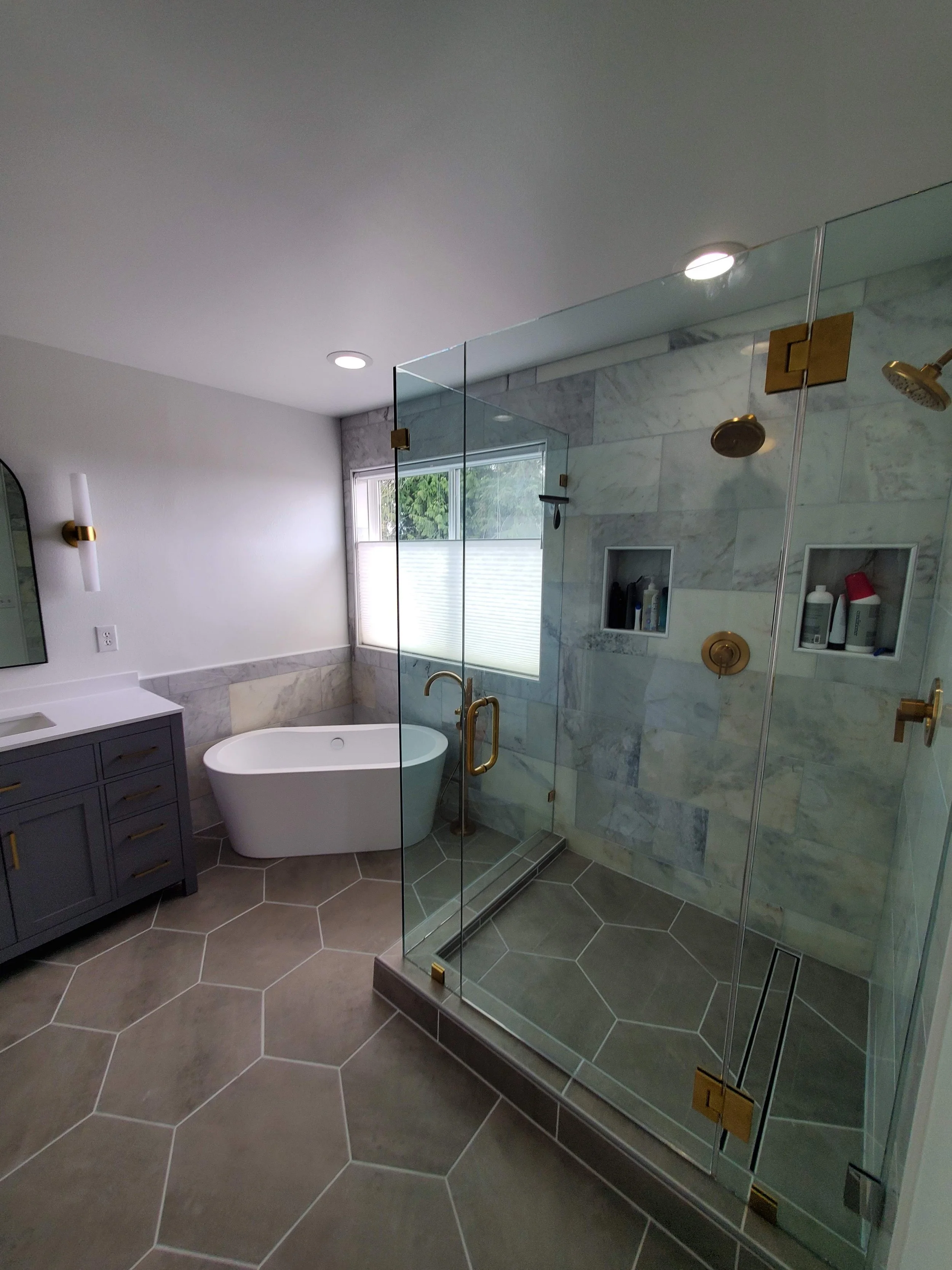 Modern bathroom with a glass shower enclosure, a white bathtub, a gray vanity with gold hardware, and hexagonal floor tiles.