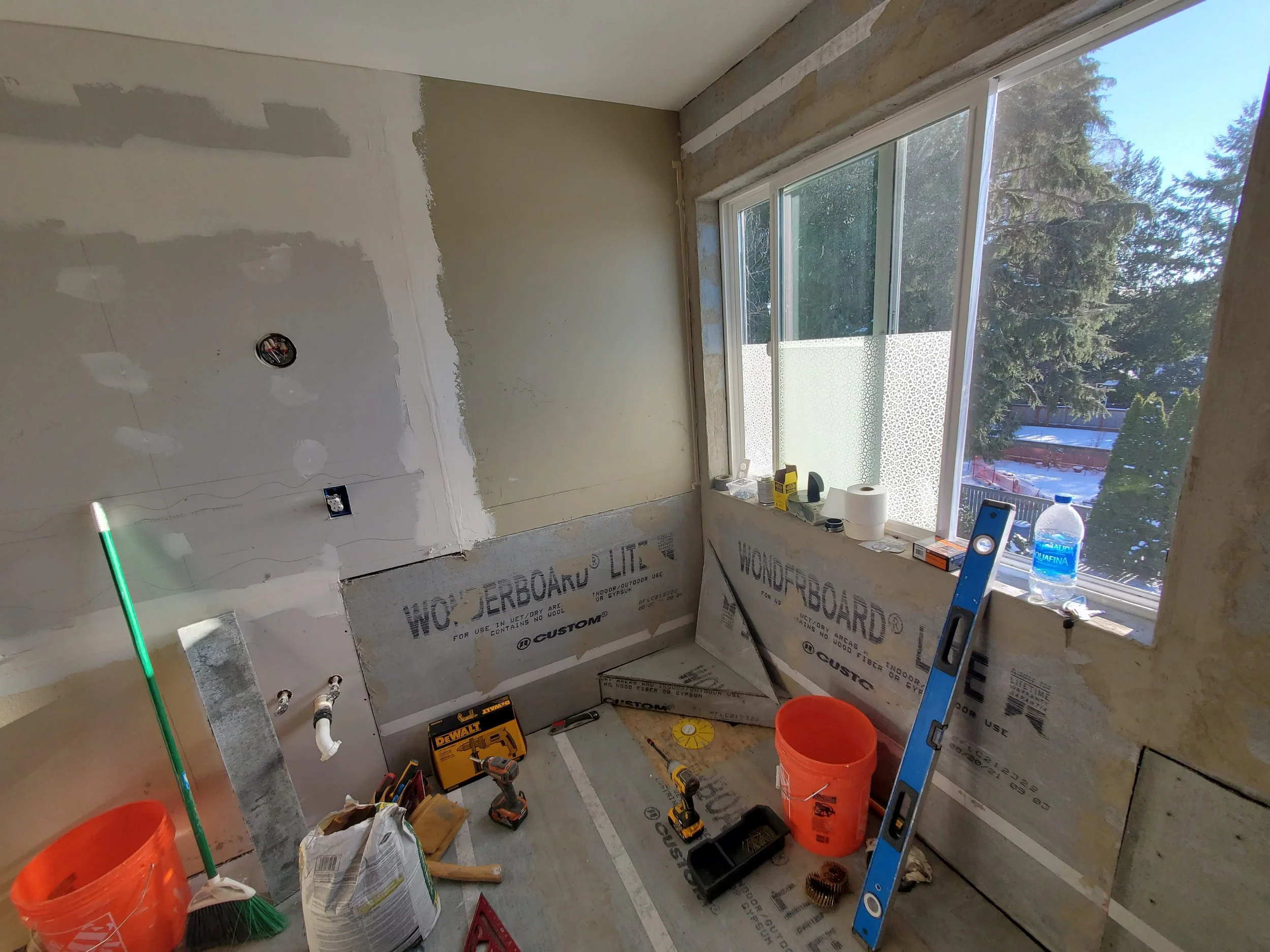 Room under construction with unfinished walls, construction tools, and materials such as an orange bucket, drill, level, vacuum, and a window overlooking trees outside.