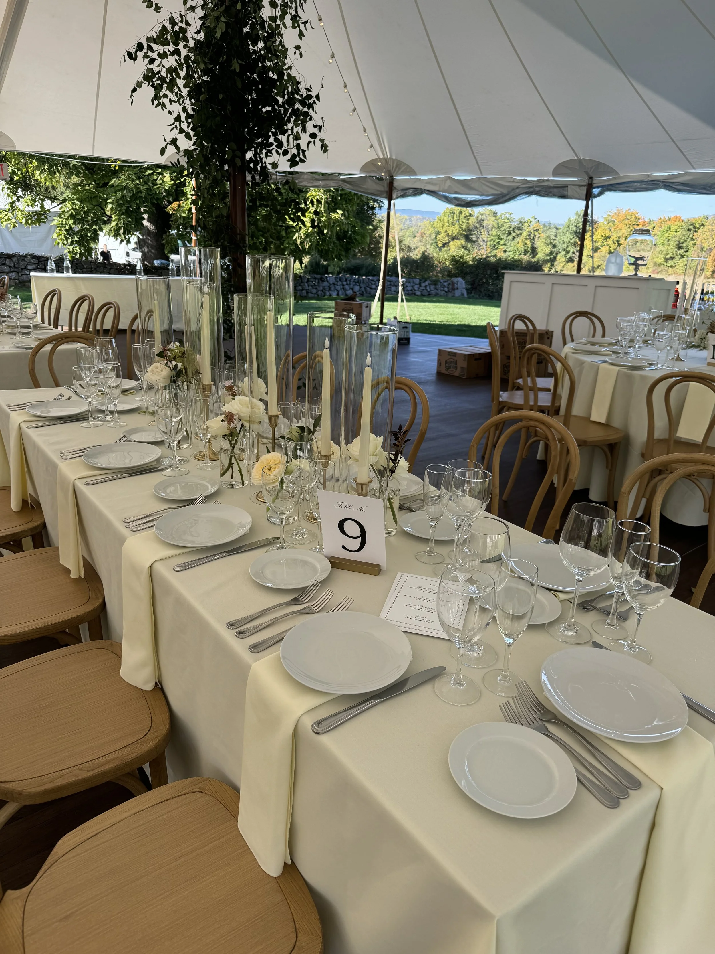 An elegant event table set with white tablecloths, white plates, silverware, wine glasses, and floral centerpieces in tall glass vases, under a large tent with trees and outdoor scenery in the background.