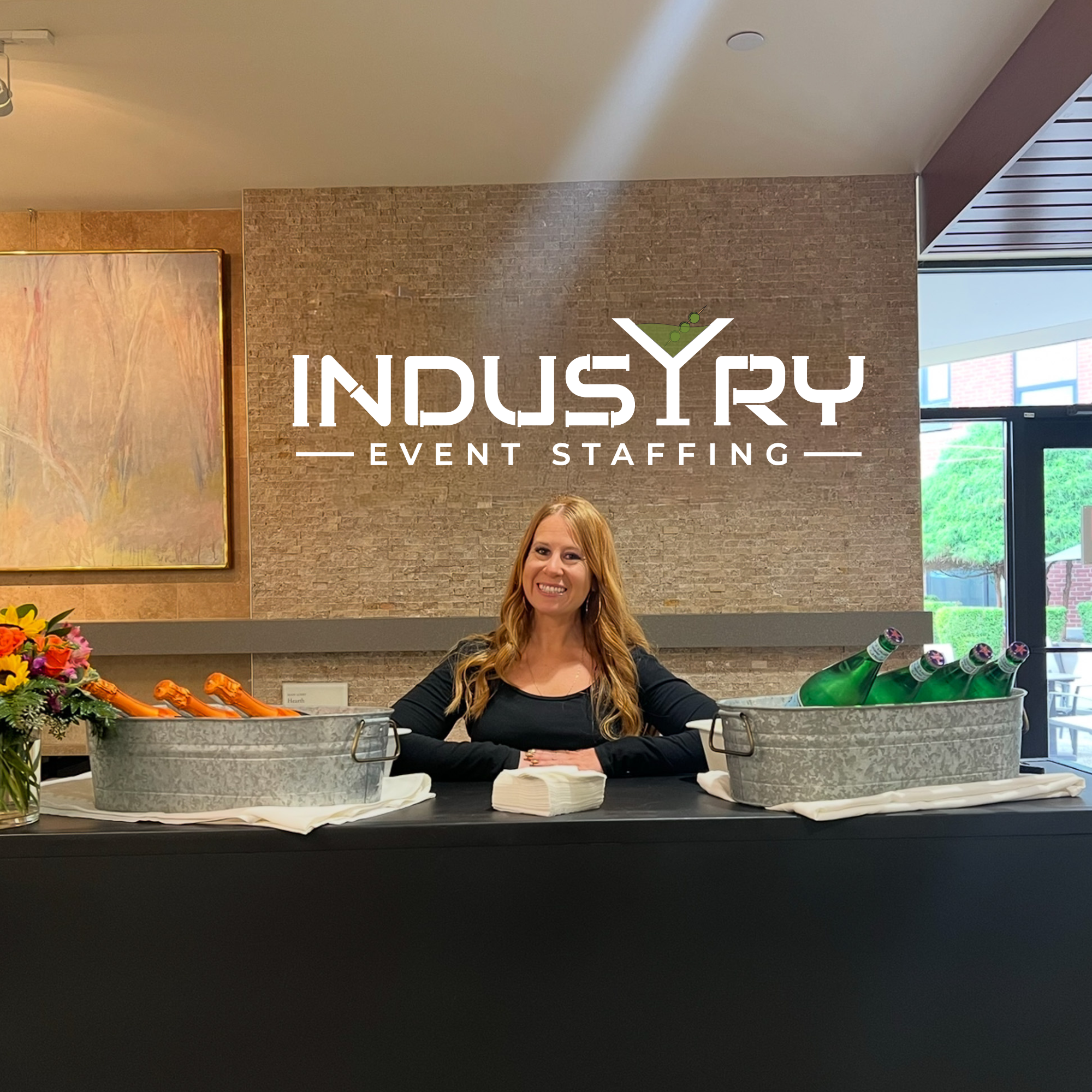 A woman with long red hair smiling at a registration desk decorated with flowers and drinks during an event at Industry Event Staffing.