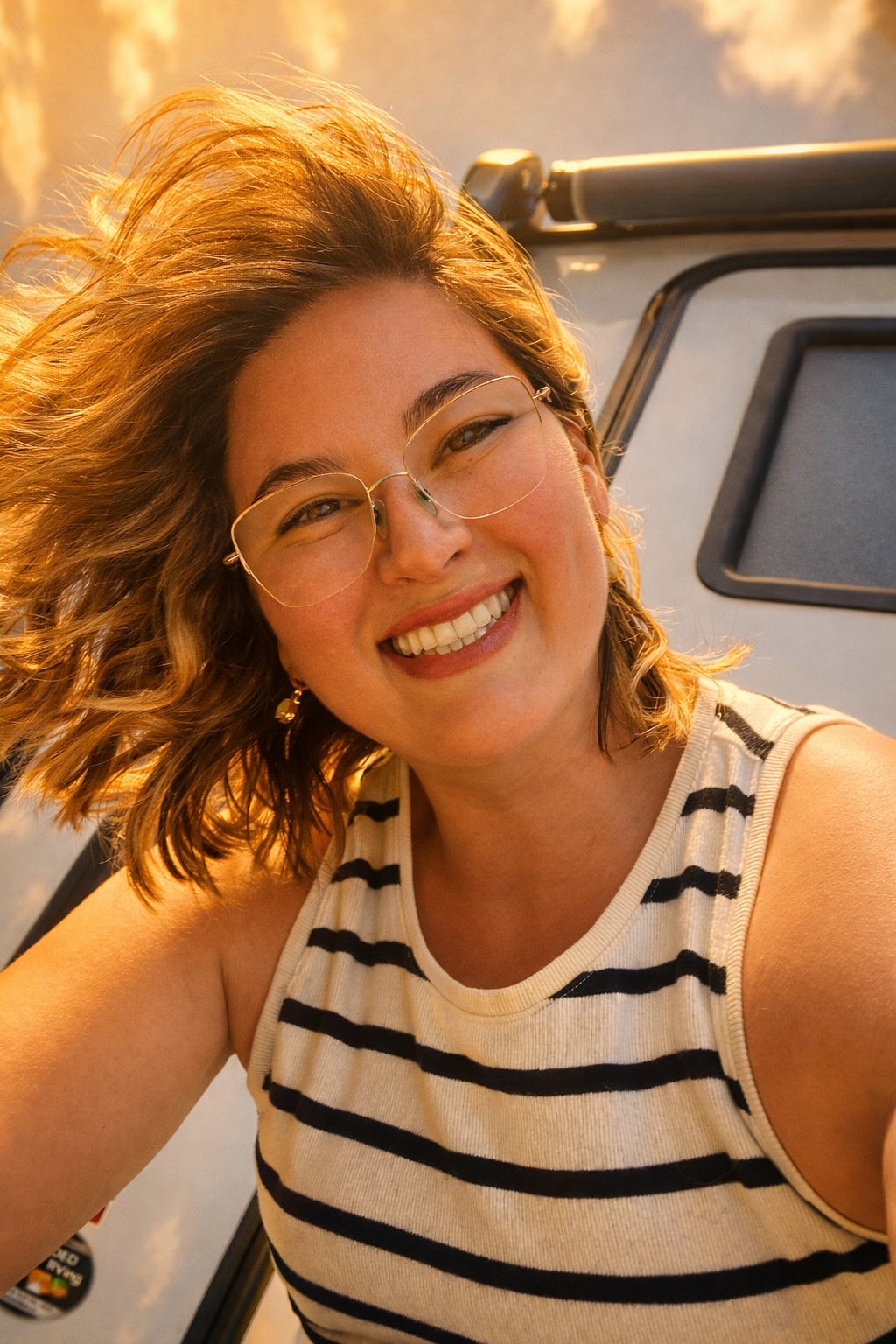 A woman with curly hair, wearing glasses and a striped sleeveless top, smiling at sunset.