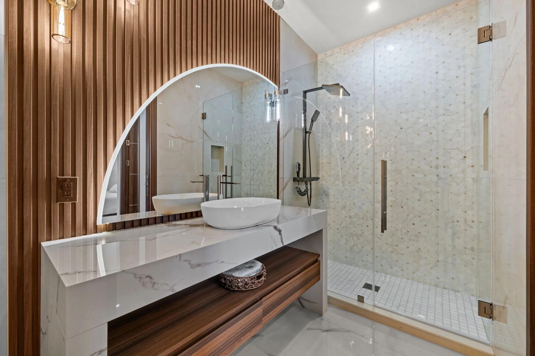 Modern bathroom with a glass-enclosed shower, a bathroom vanity with a white marble countertop, a vessel sink, and wooden paneling. The shower has a black rainfall showerhead and handheld shower, with beige patterned tiles on the walls.