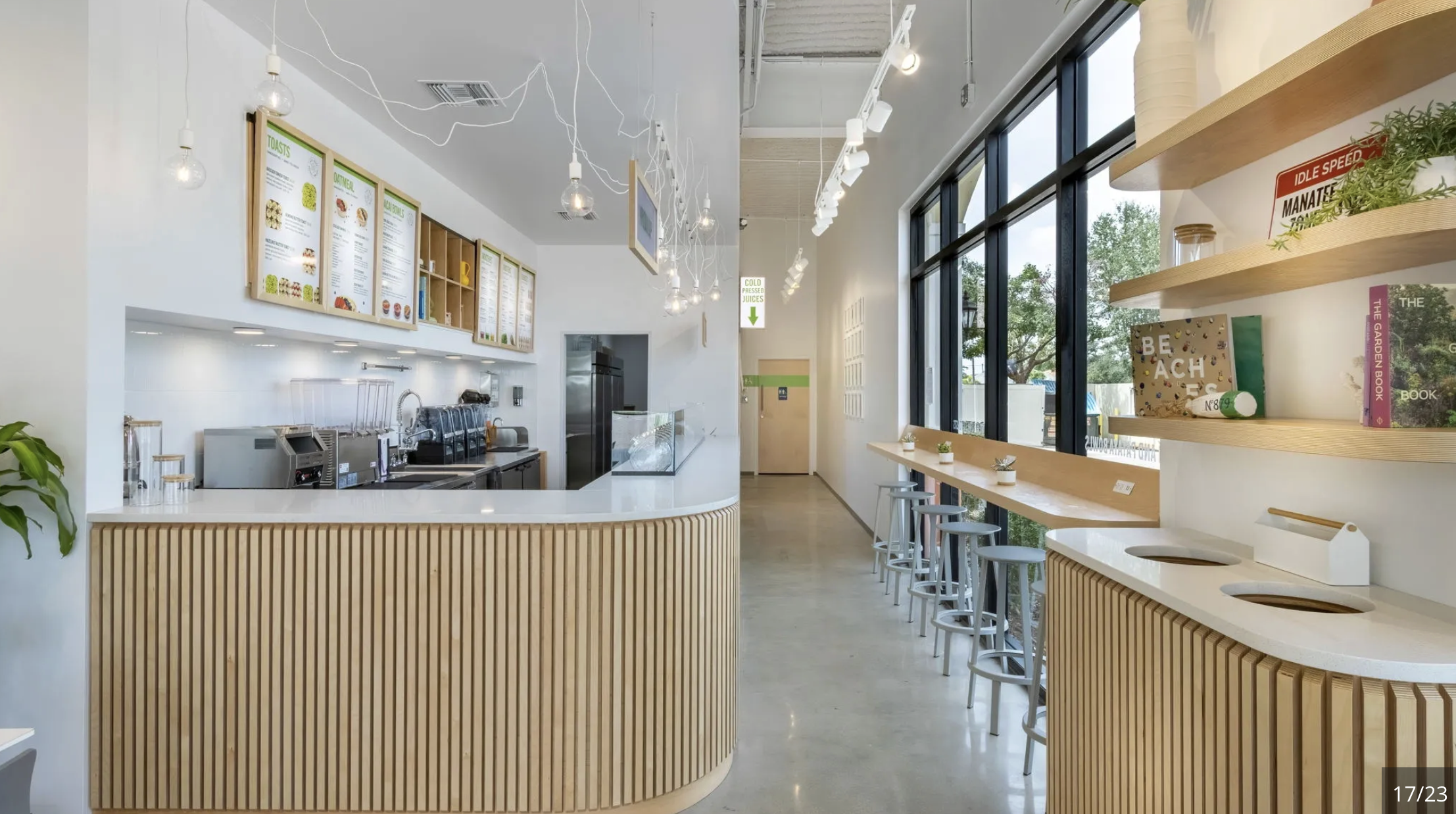 Interior of a modern, minimalist cafe with a white counter, wooden accents, and barstools along a large window with greenery outside.