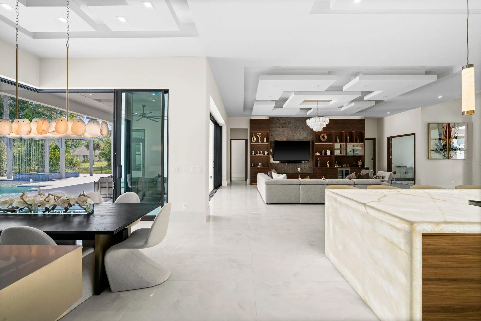 Modern open-concept living room with a large sectional sofa, built-in wooden shelves, and a flat-screen TV, with sliding glass doors leading outside to a pool and patio area.