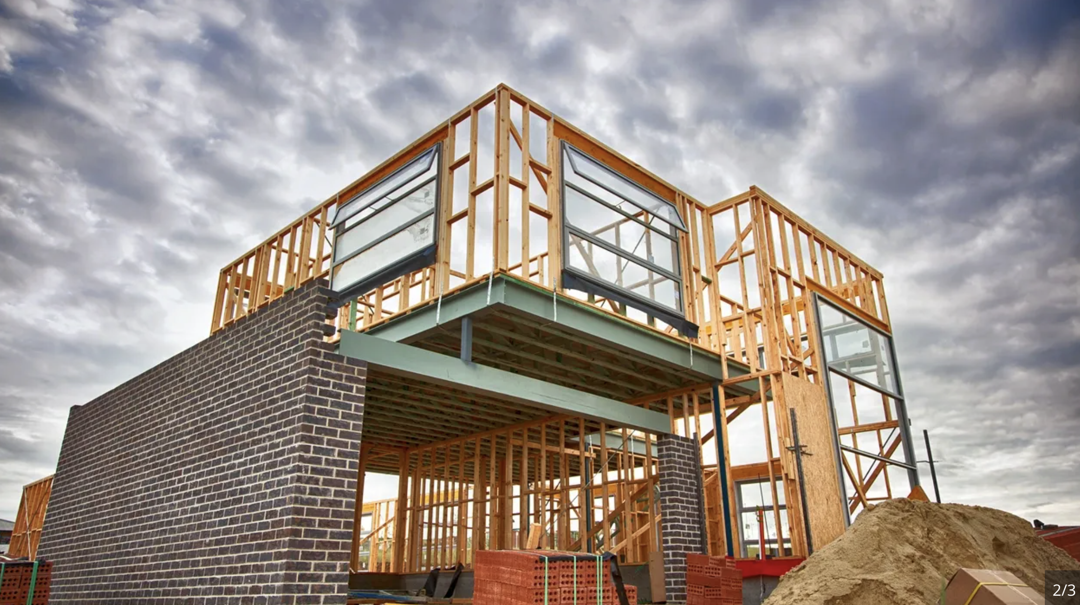 Construction site with a building under construction, showing wooden framing and a brick wall base, with cloudy sky overhead.
