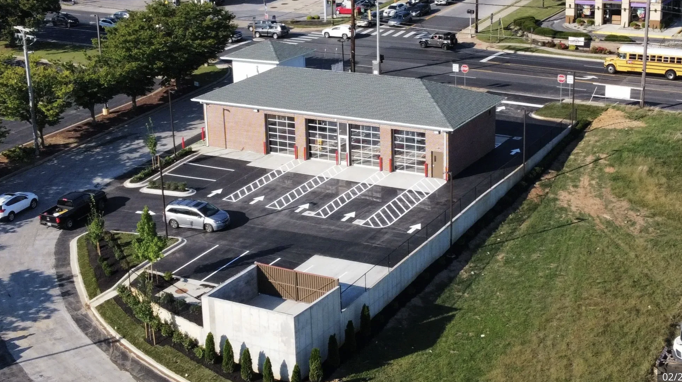 An aerial view of a small fire station building with three garage doors, surrounded by a parking lot with a few parked cars. The building is flanked by green trees and landscaped areas, with roads and traffic visible in the background.