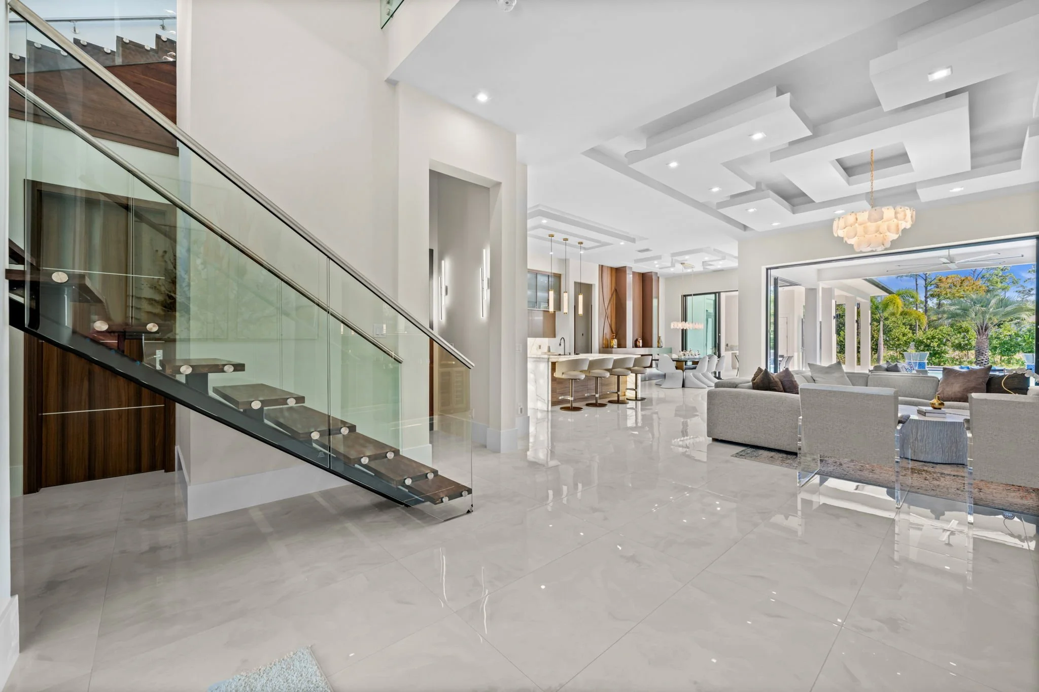 Modern open-concept living room and kitchen with white walls, high ceilings, luxurious furniture, and large windows showing outdoor greenery.