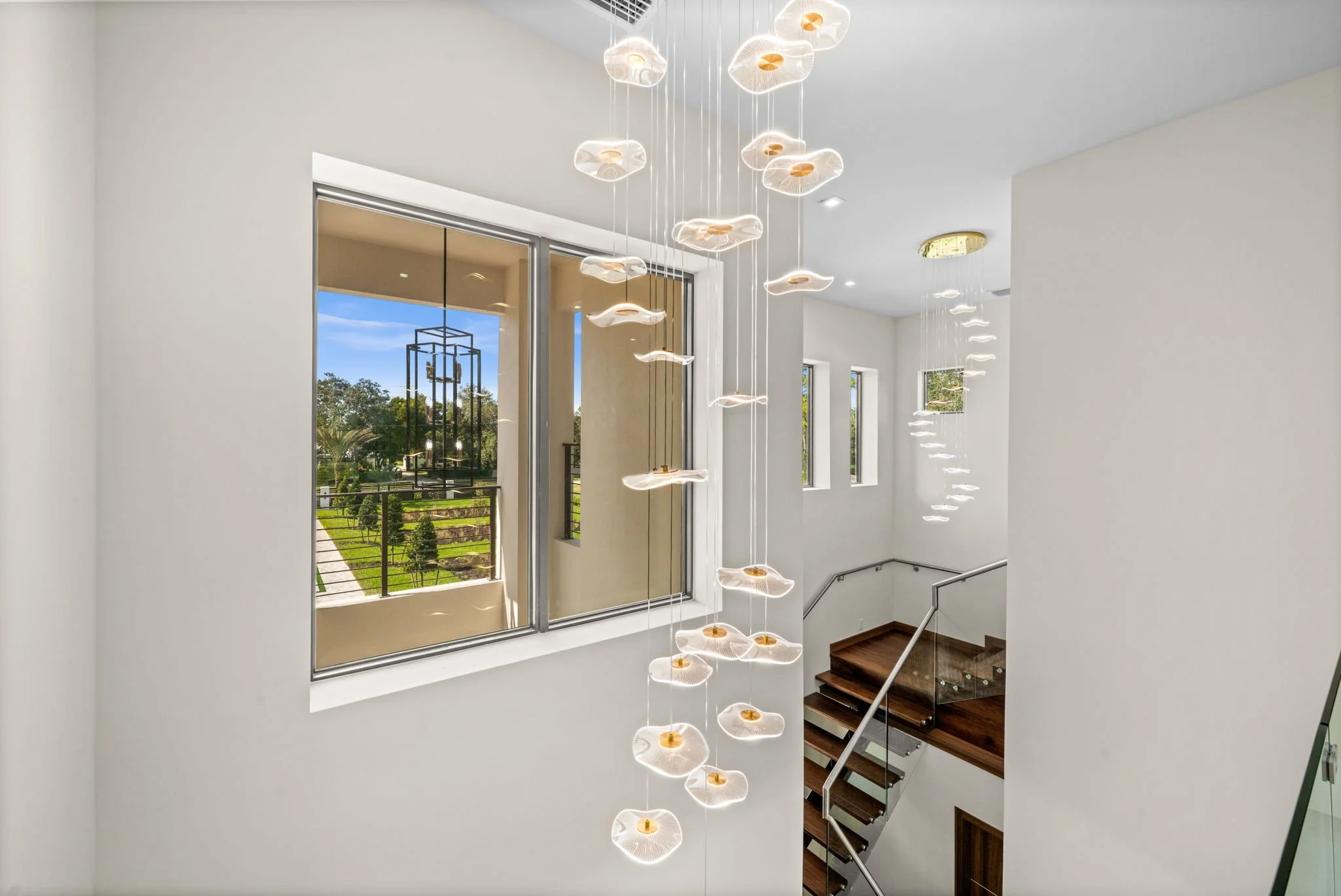 Modern interior staircase area with large window, hanging contemporary chandelier, and sleek railing.