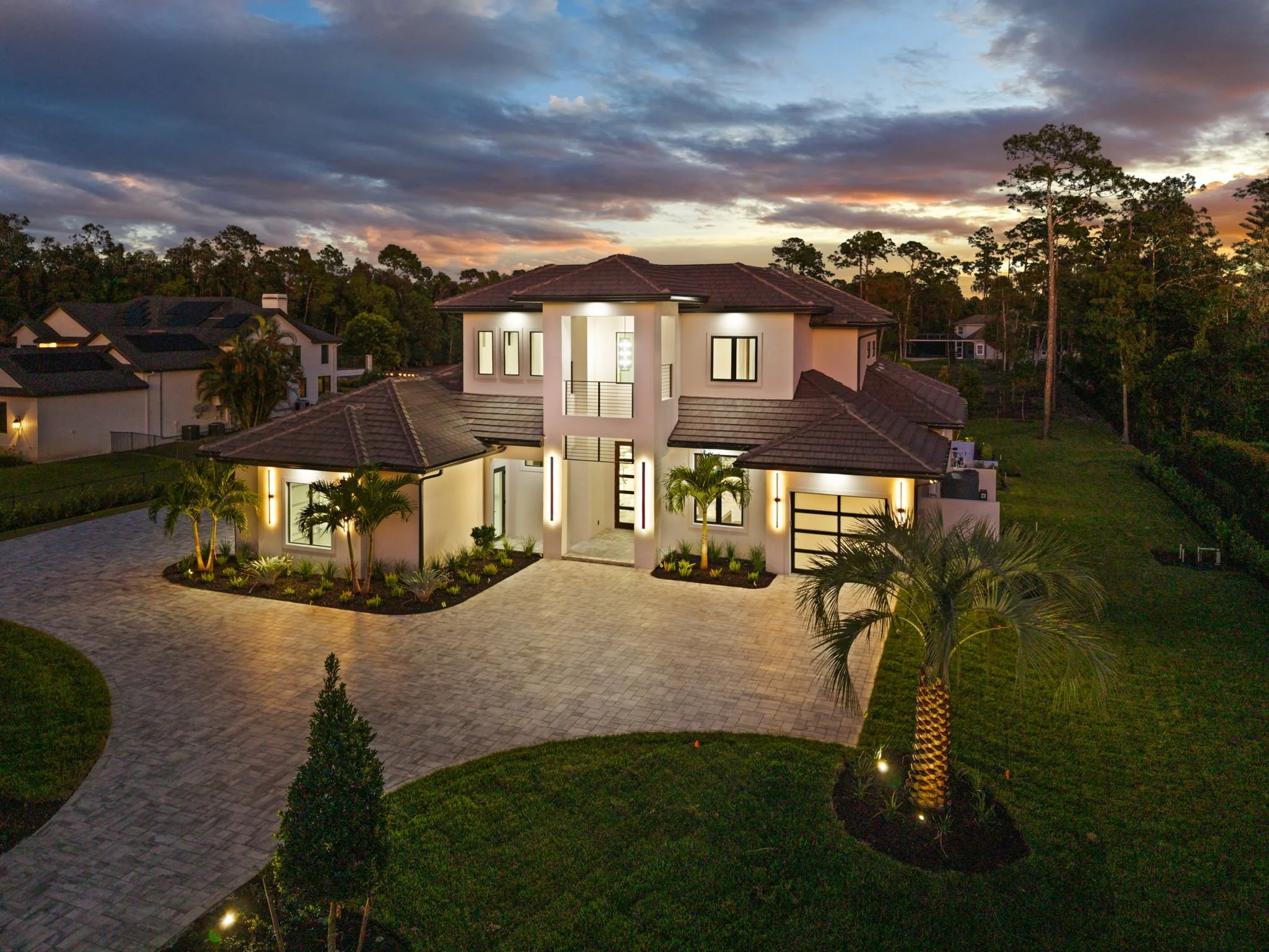 A modern residential house at sunset with illuminated exterior lighting, a circular paved driveway, and landscaped yard with palm trees and plants. The sky has clouds with a colorful sunset glow.