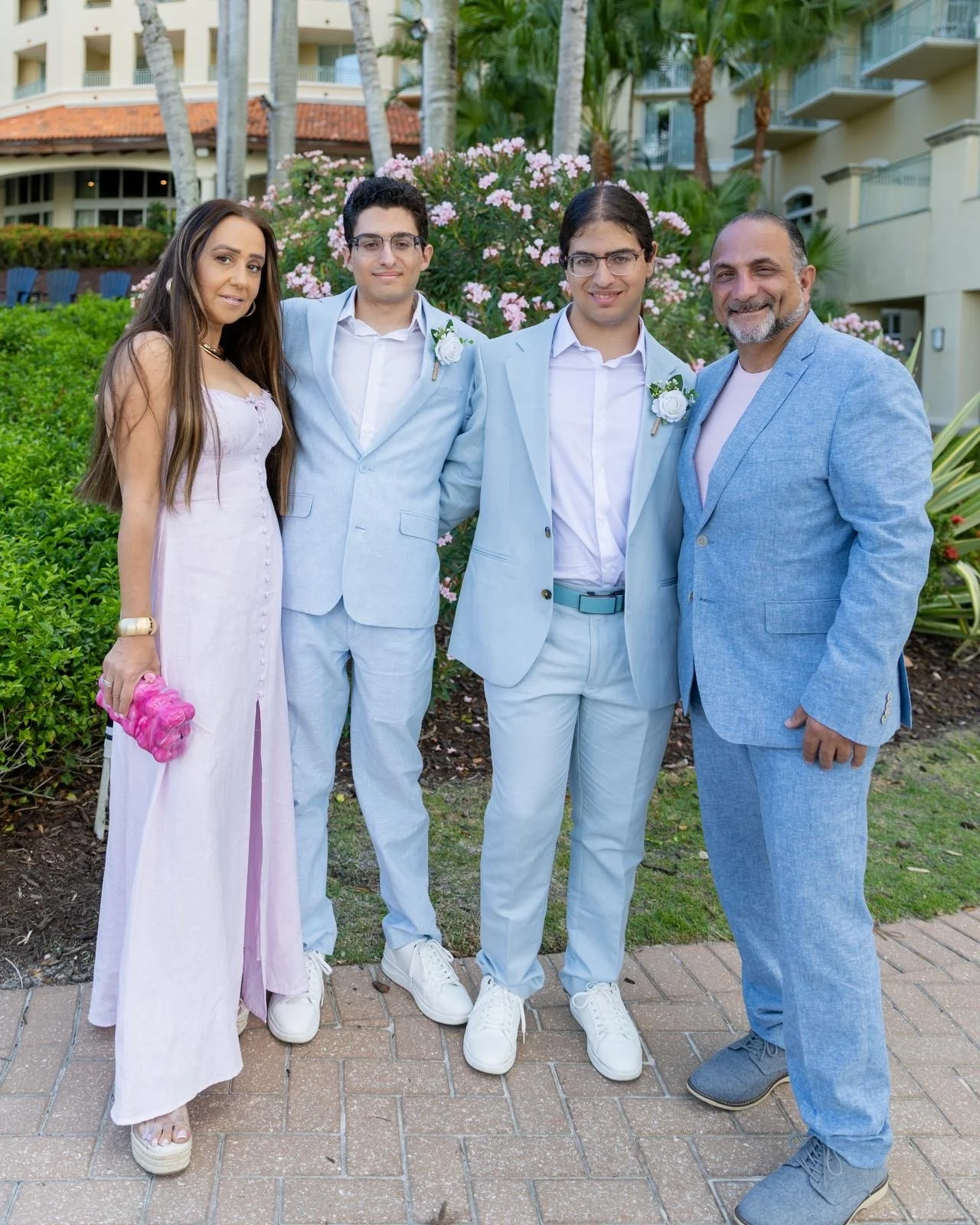 A group of five people, including two men and one woman, standing outdoors in front of pink flowering bushes and a building with balconies. They are dressed in light-colored, semi-formal attire, suggesting a celebratory event like a wedding or party.