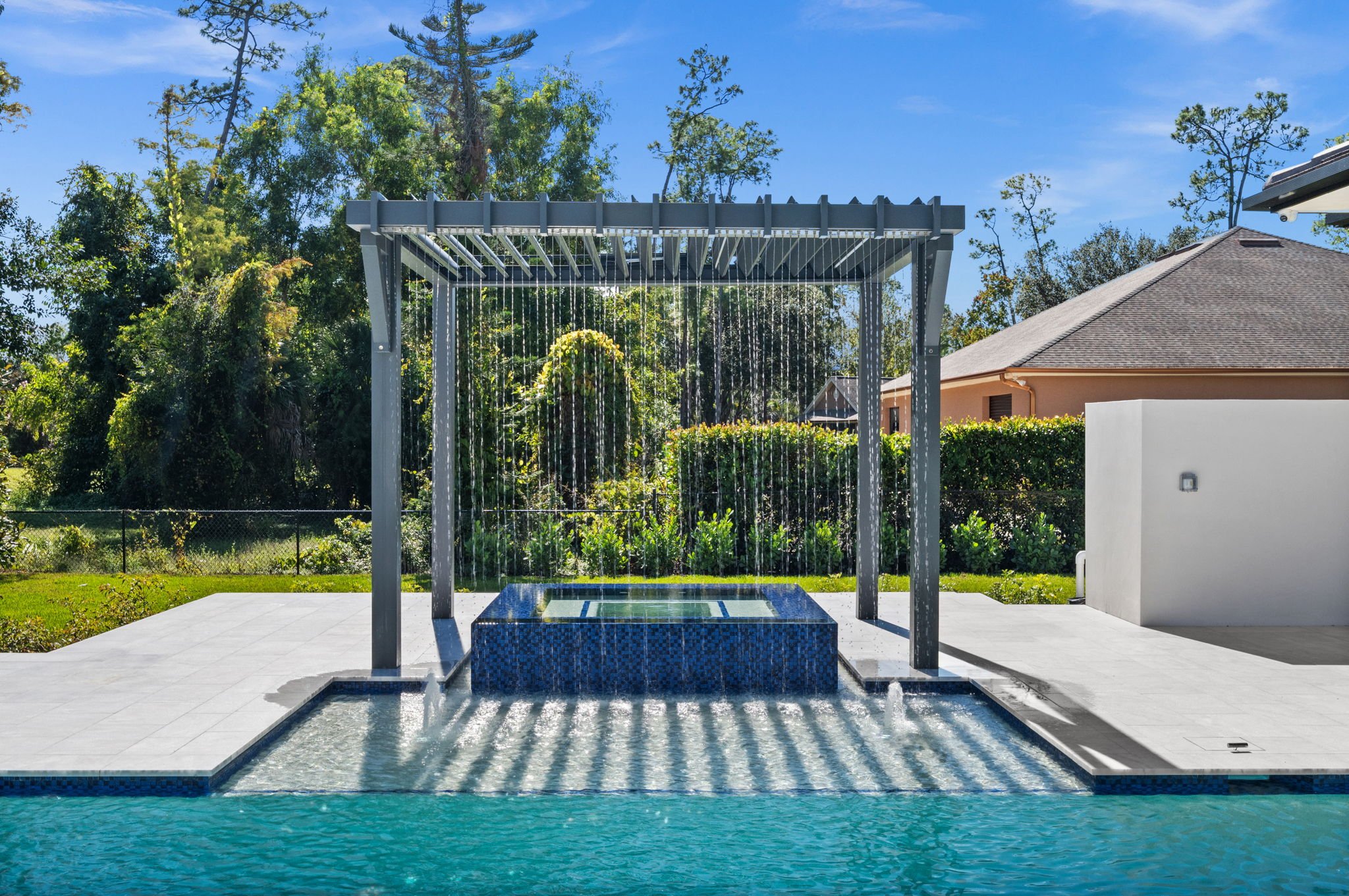 An outdoor shower structure with a waterfall feature, located near a swimming pool, with trees and a house in the background.