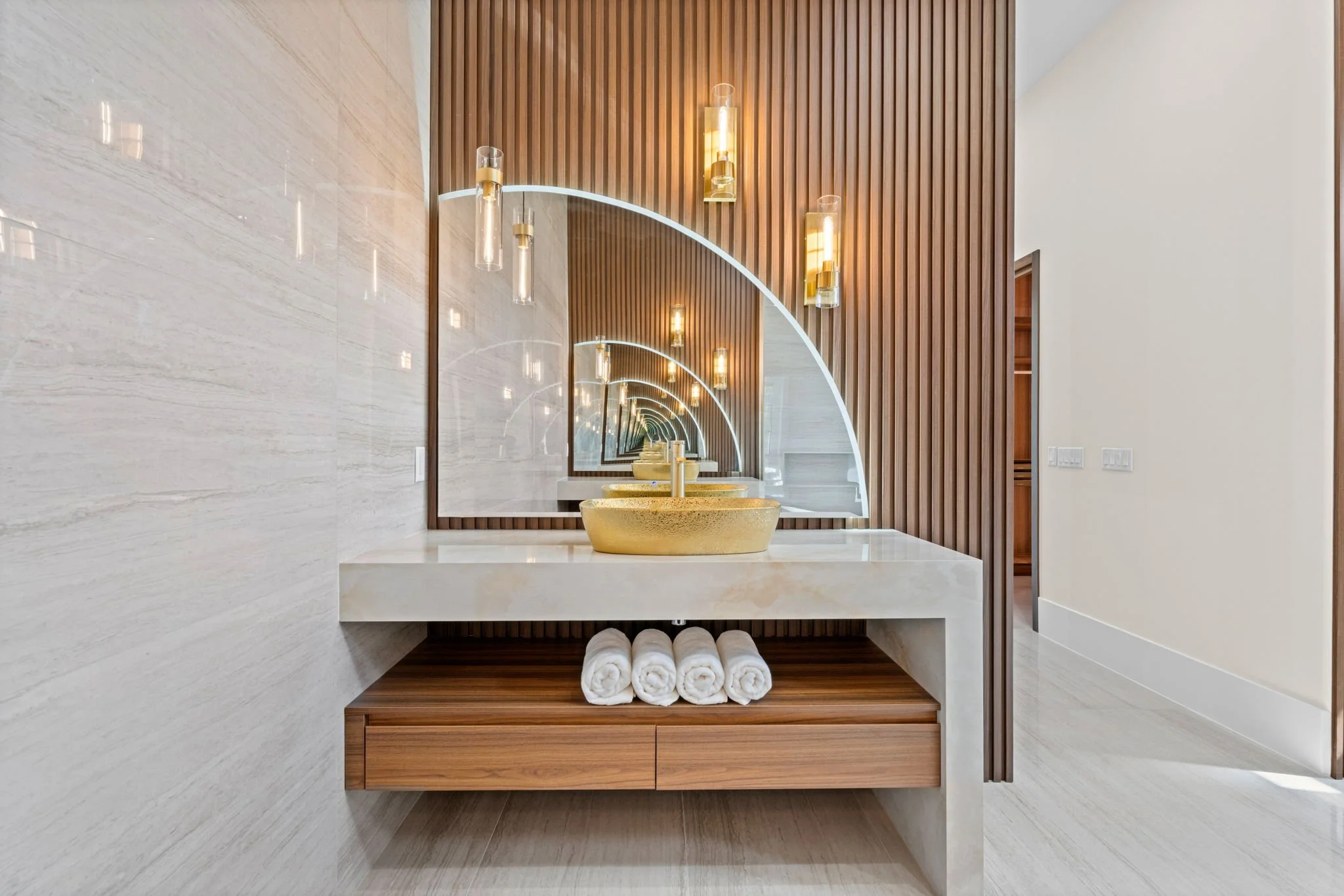 Modern bathroom vanity with a marble countertop, gold sink bowls, four rolled white towels, and a large mirror with pendant lights against a wood-paneled wall.