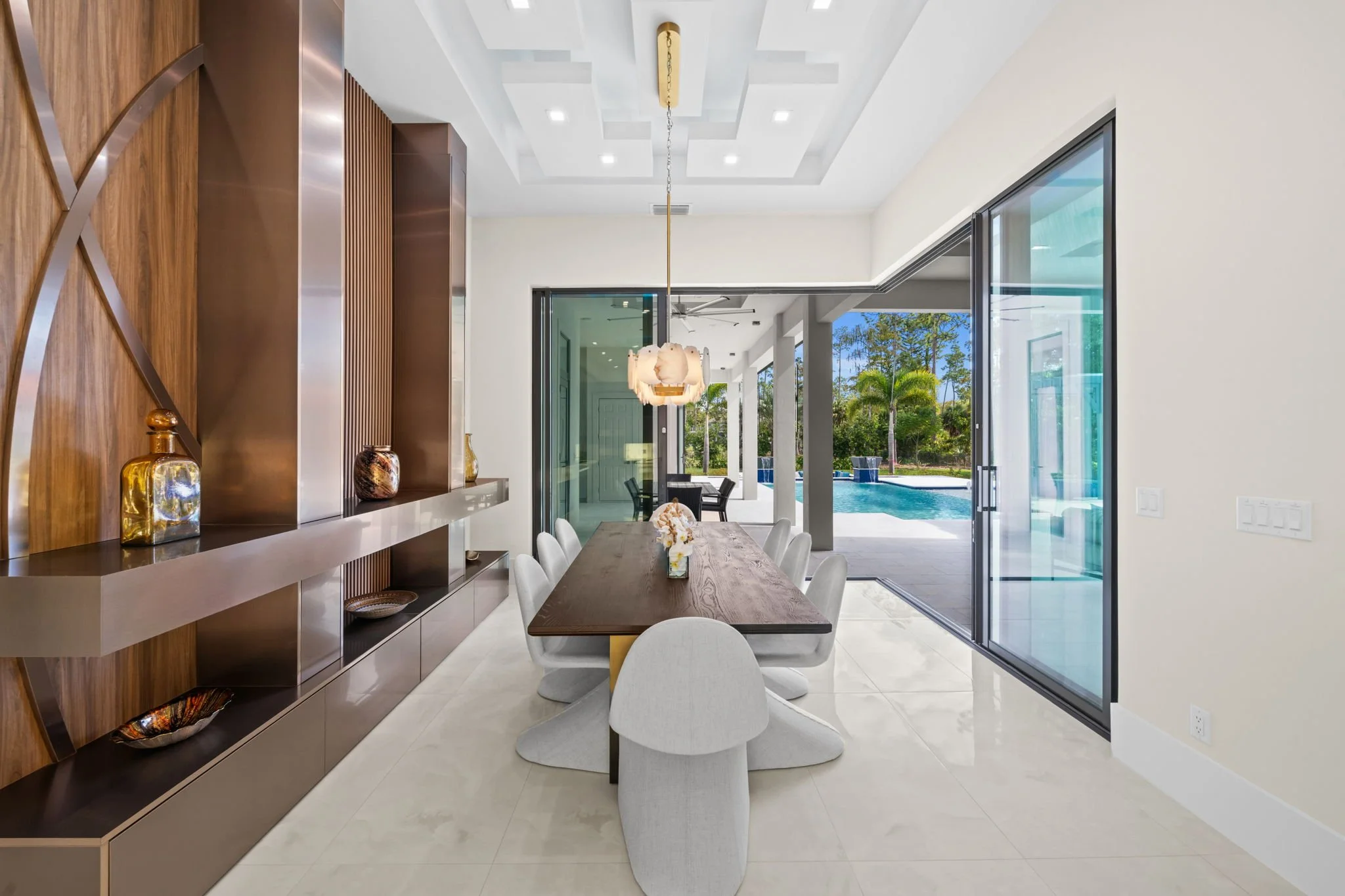 Modern dining area with a wooden dining table, white cushioned chairs, and a decorative light fixture hanging over the table. Large sliding glass doors reveal a backyard with a pool and trees outside.