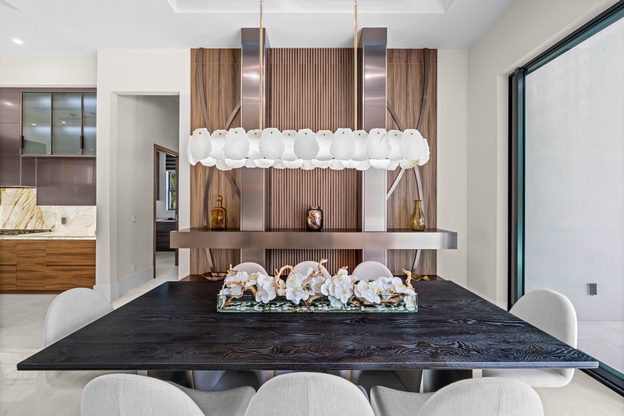 Modern dining area with black wooden table, white upholstered chairs, floral centerpiece, wooden and metal wall decor, and a pendant light fixture with cloud-like white shades.