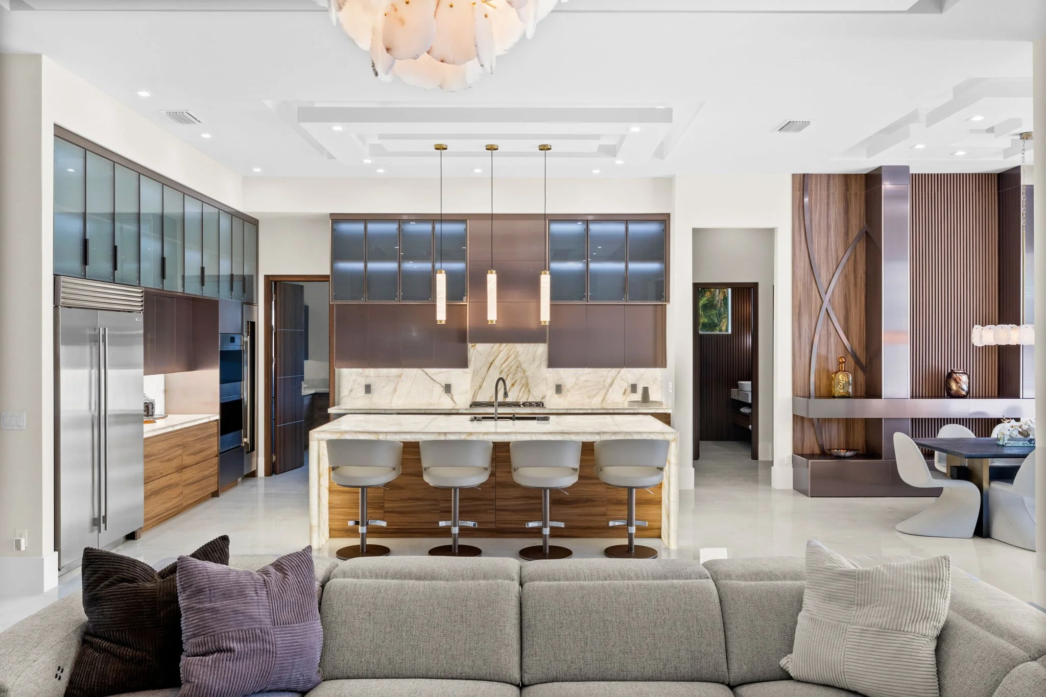 Modern open-concept kitchen and living room with white walls and ceiling, marble countertop island, bar stools, wooden cabinetry, shelving, pendant lighting, and a sectional sofa with purple cushions in the living area.