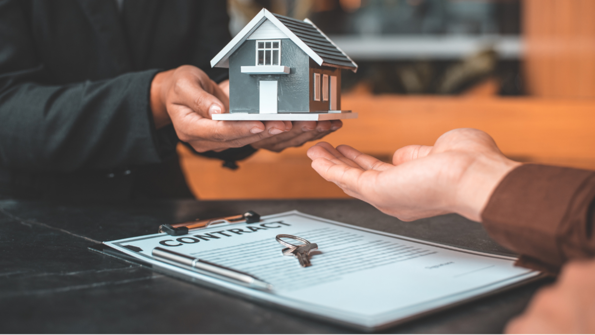 A person is handing over a small model house to another person at a table with a contract and keys.