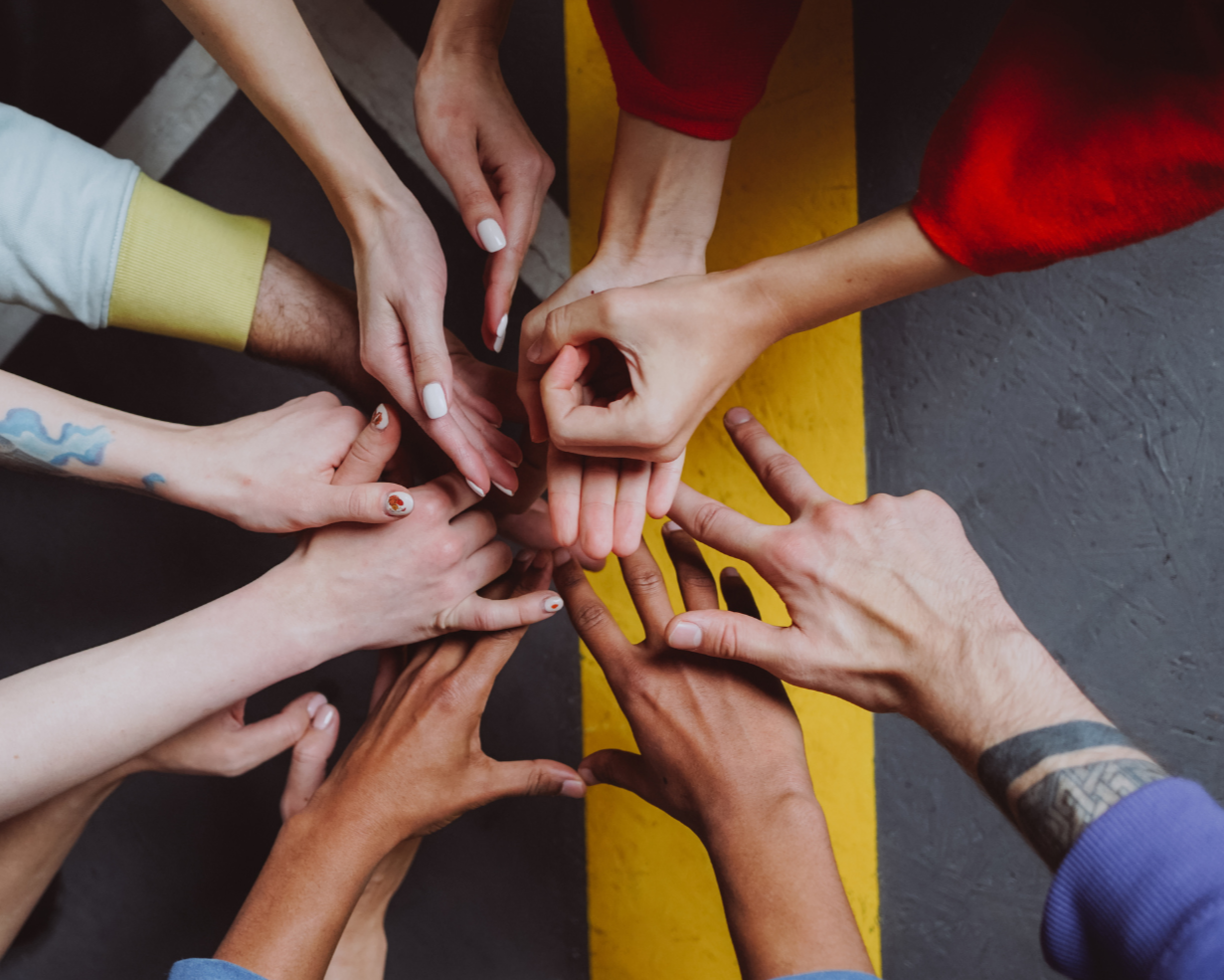 Group of diverse hands reaching towards the center, forming a circle on a concrete surface marked with a yellow line.