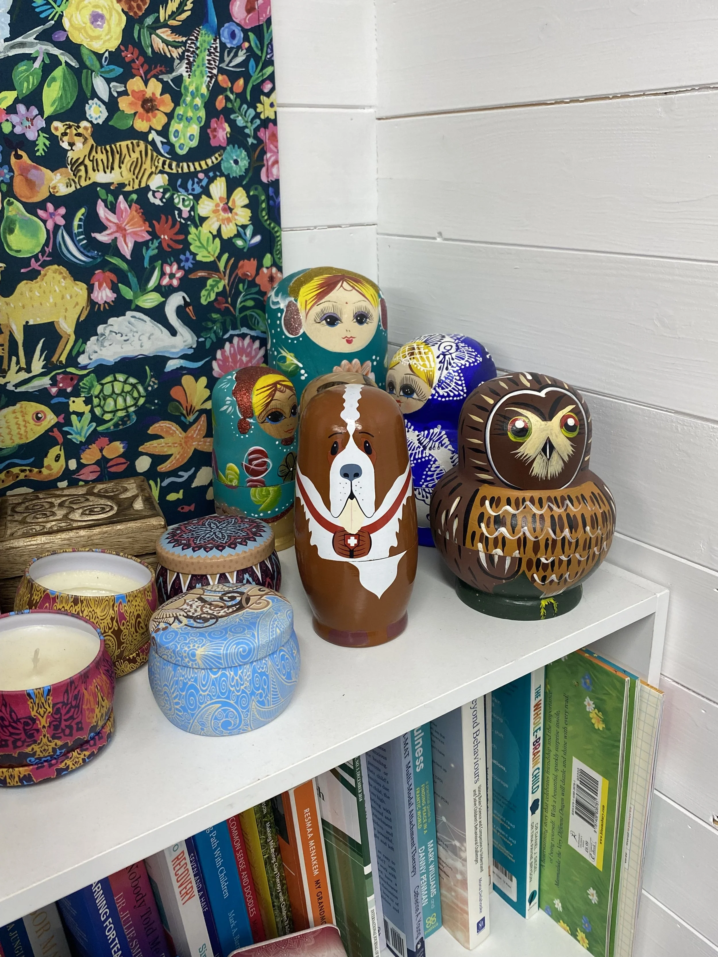 Colorful decorative Russian nesting dolls, candles, and carved wooden animal figures on a white shelf, with a vibrant floral and animal patterned cloth in the background.