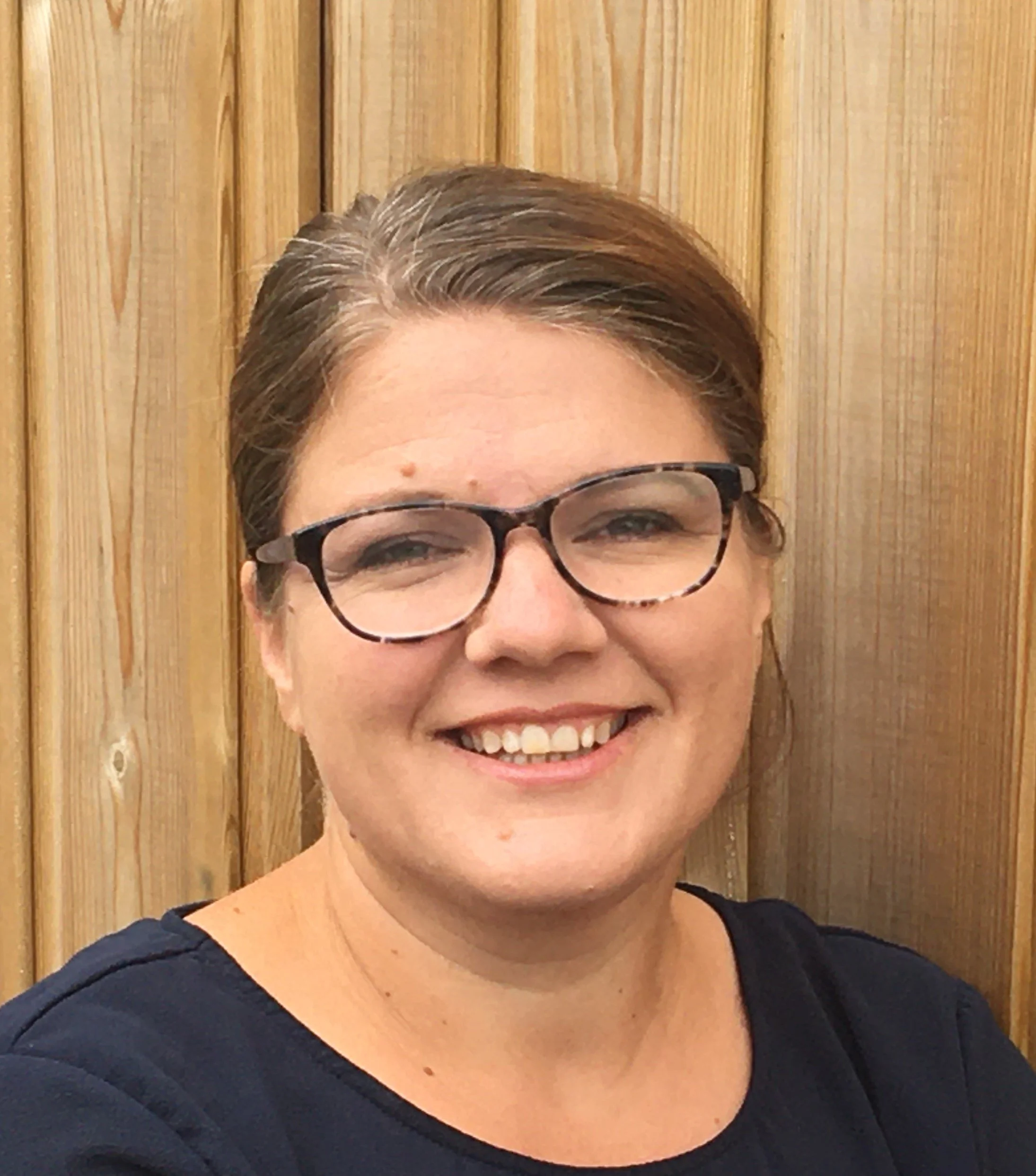 A woman with short brown hair, glasses, and a big smile standing in front of a wooden wall.