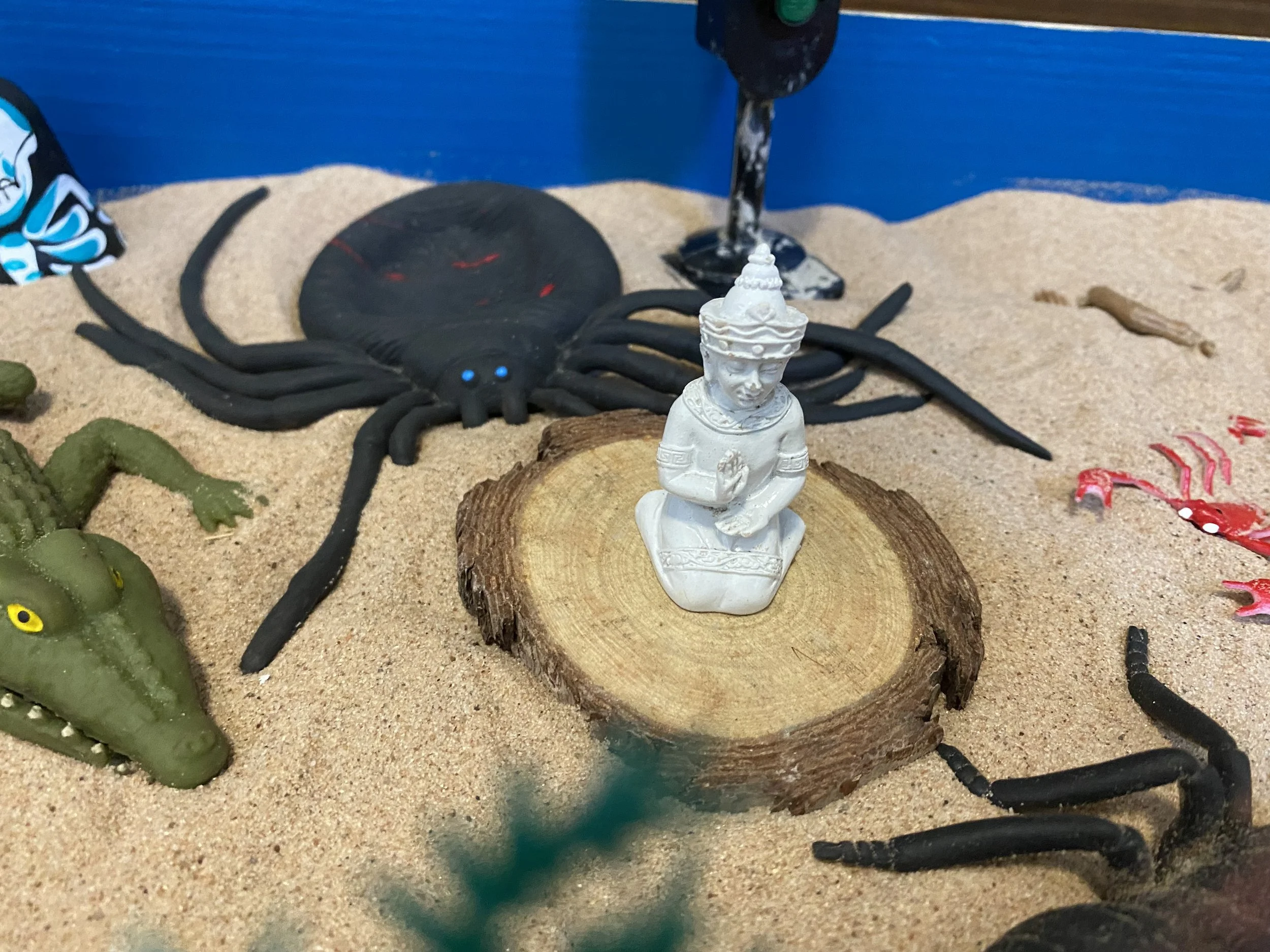 Miniature desert scene with toy alligator, a large toy spider, a small figurine of a person in traditional attire kneeling in prayer, and various small plastic insects on sand with a blue background.