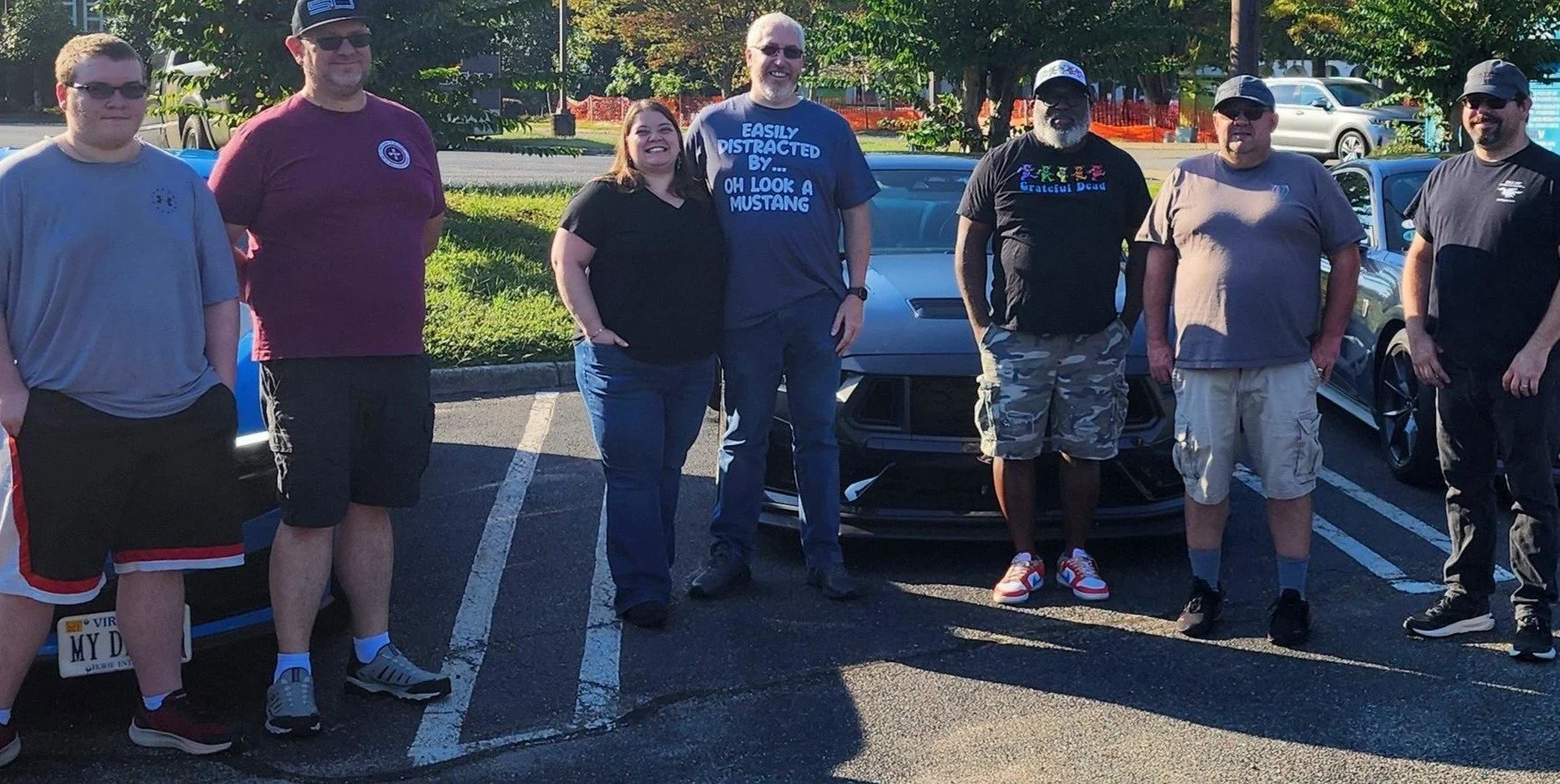 Group of people standing in a parking lot, some in front of a collection of Ford Mustangs, outdoors with green trees in the background, smiling and posing for a photo.