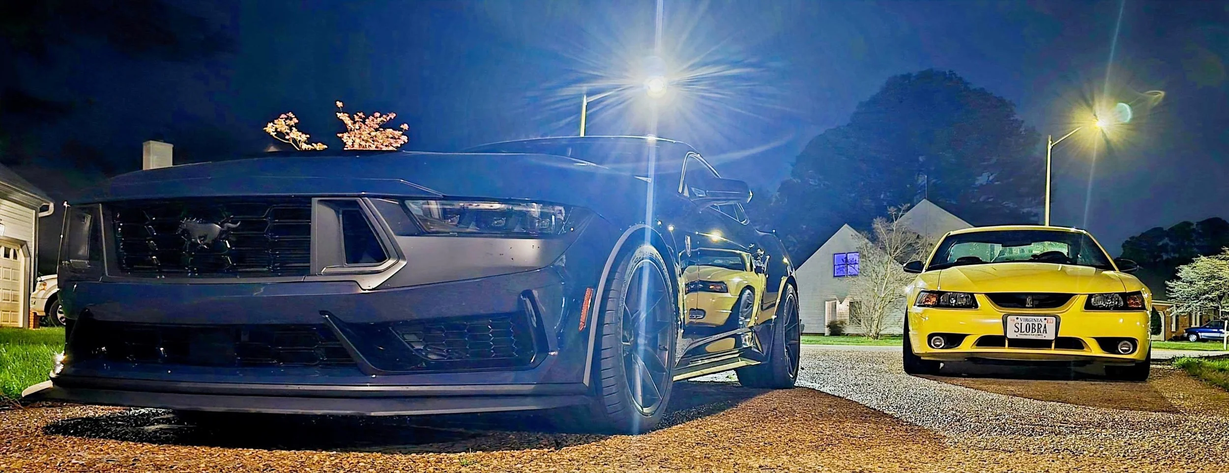 Nighttime view of a yellow Ford Cobra and Blue Ember Metallic Ford Mustang Dark Horse parked on a suburban street, illuminated by streetlights and surrounded by houses and trees.