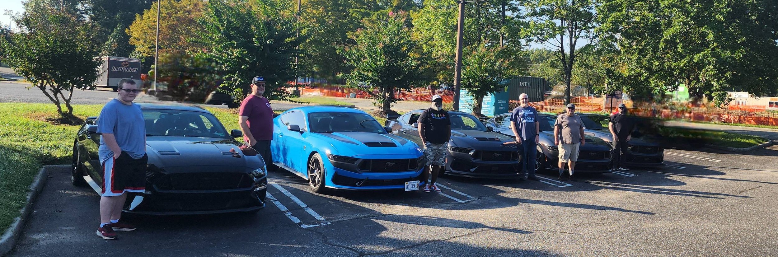 A group of seven people standing in front of six black and blue Ford Mustang cars parked in a parking lot with trees and a sidewalk in the background.