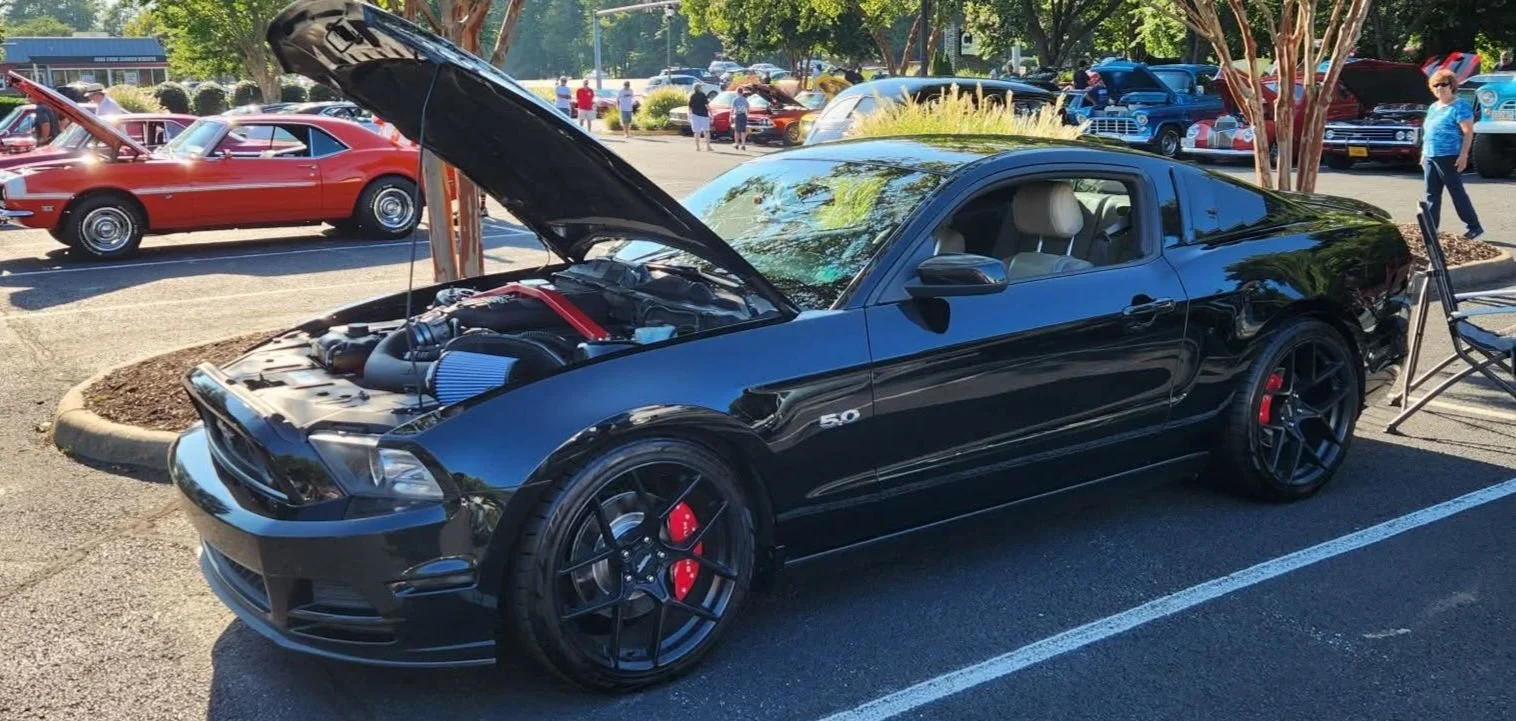 A black Ford Mustang with its hood open at a car show, showcasing its engine. The car has black wheels with red brake calipers. Other classic and vintage cars are visible in the background, and people are walking around.
