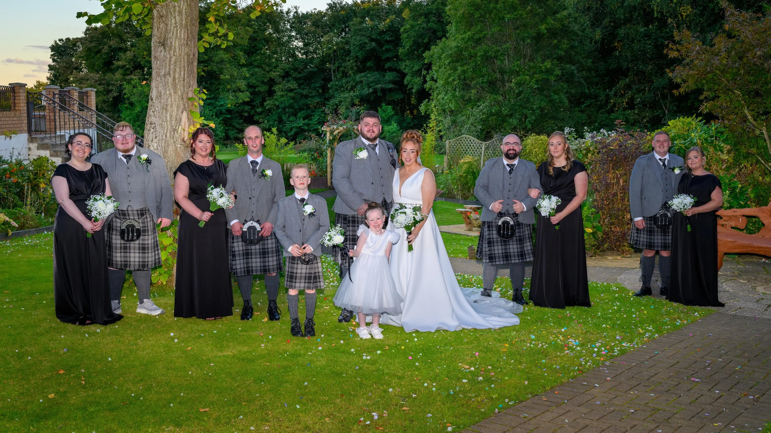 A wedding party of twelve people, including the bride and groom, standing on a lawn with scattered confetti, greenery, and trees in the background, during evening.