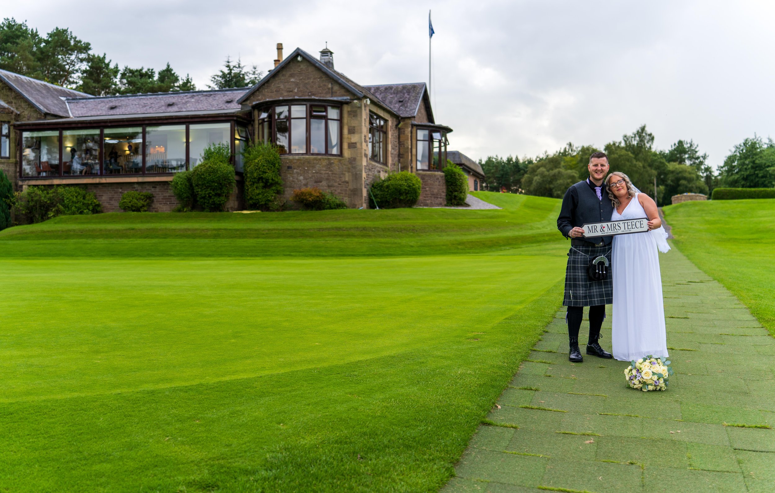 A newlywed couple stands on a green path outside a large stone house, holding a sign that reads 'Mr & Mrs Teece,' with the bride in a white gown and the groom in a traditional Scottish kilt and black shirt, smiling under an overcast sky.