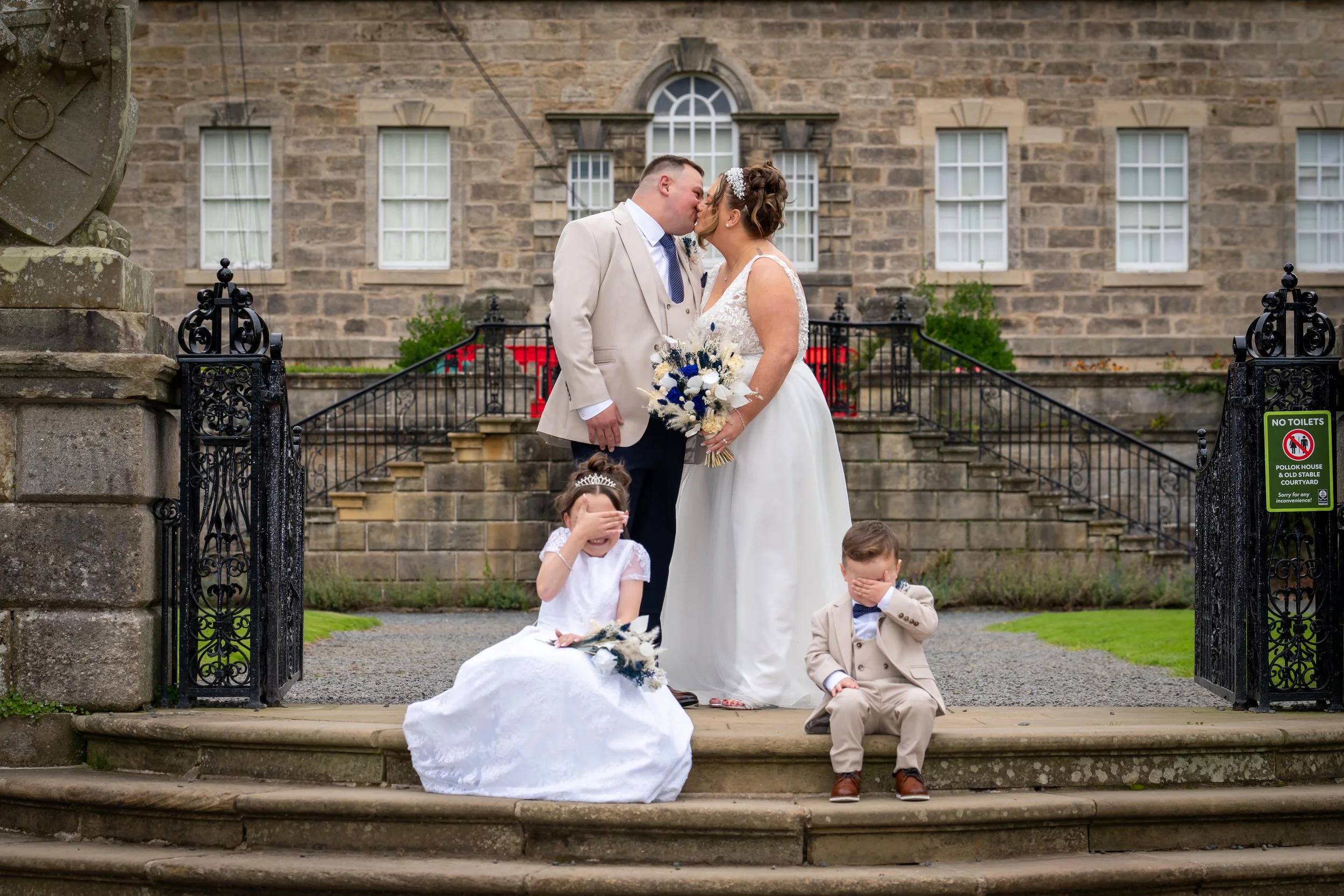 A wedding scene with a bride and groom kissing on steps in front of a brick building, with two children sitting nearby, covering their eyes.