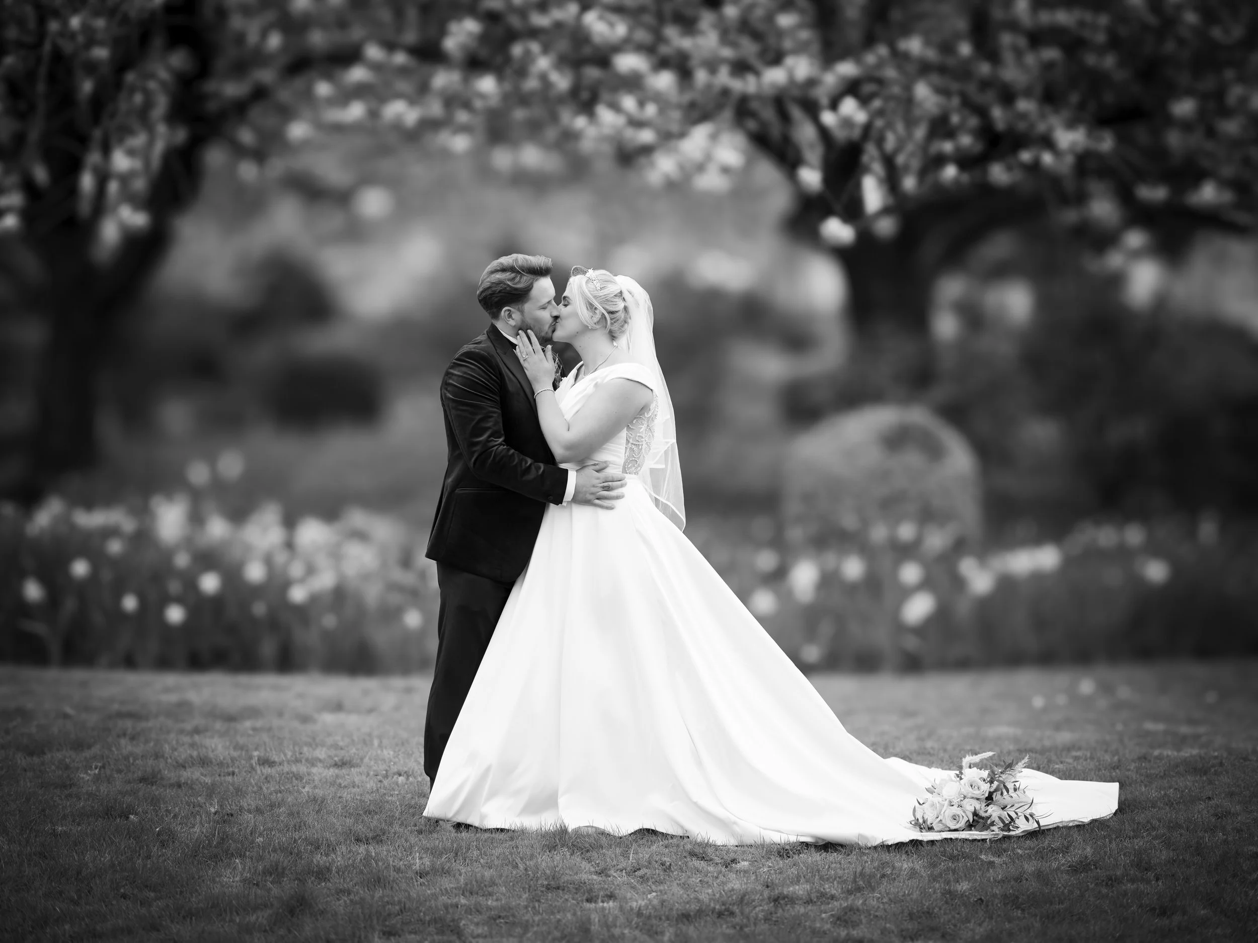 A black and white photo of a bride and groom kissing outdoors, with trees in the background and a bouquet on the ground.