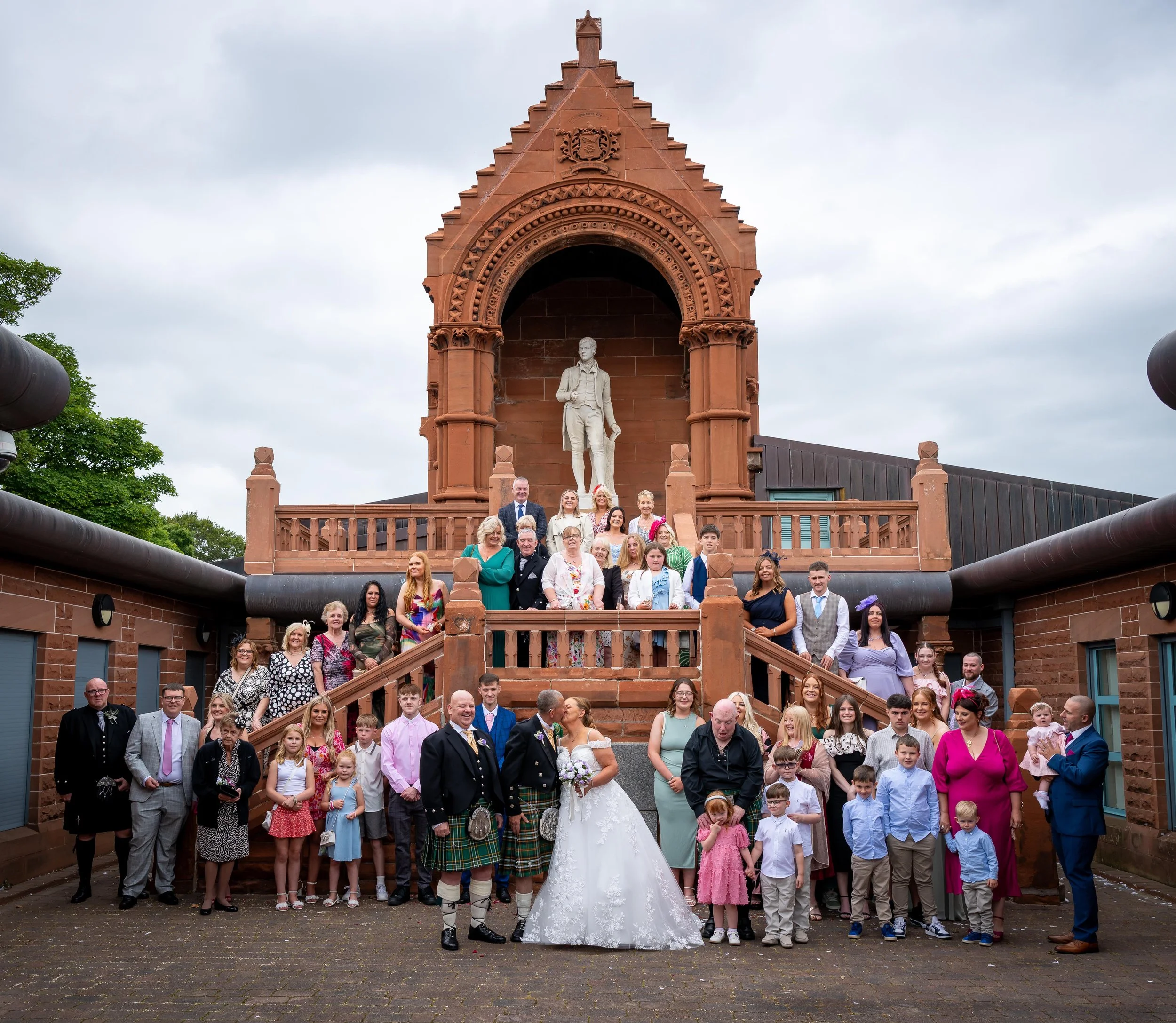 A large group of people celebrating a wedding outdoors in front of a historic red sandstone monument with a statue and clock tower. The bride and groom are in the center, hugging and surrounded by family and friends of all ages.