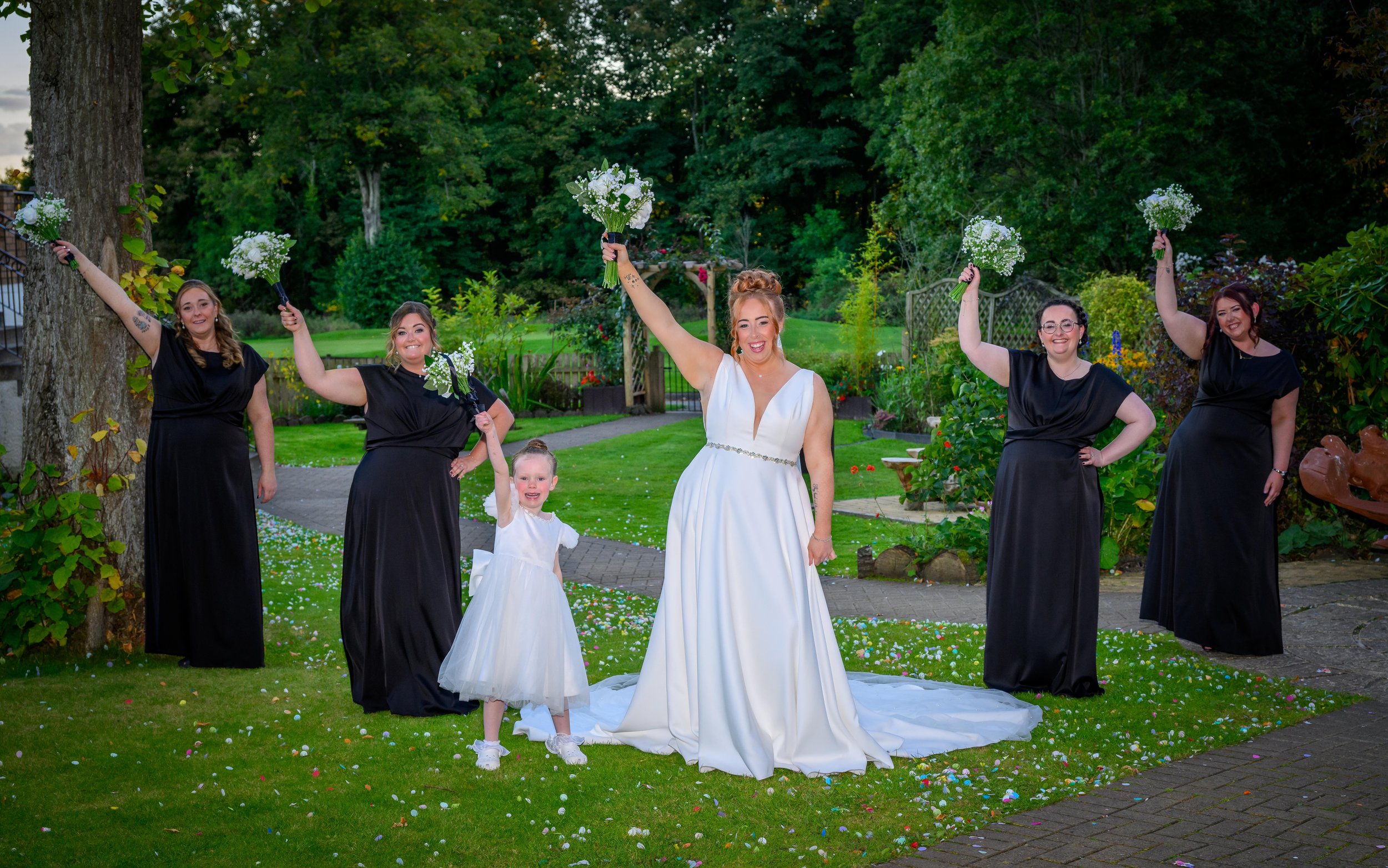 A bride in a white wedding dress stands in a garden, surrounded by five women holding bouquets, with a young girl in a white dress. They are celebrating outdoors with trees, grass, and flowers in the background.