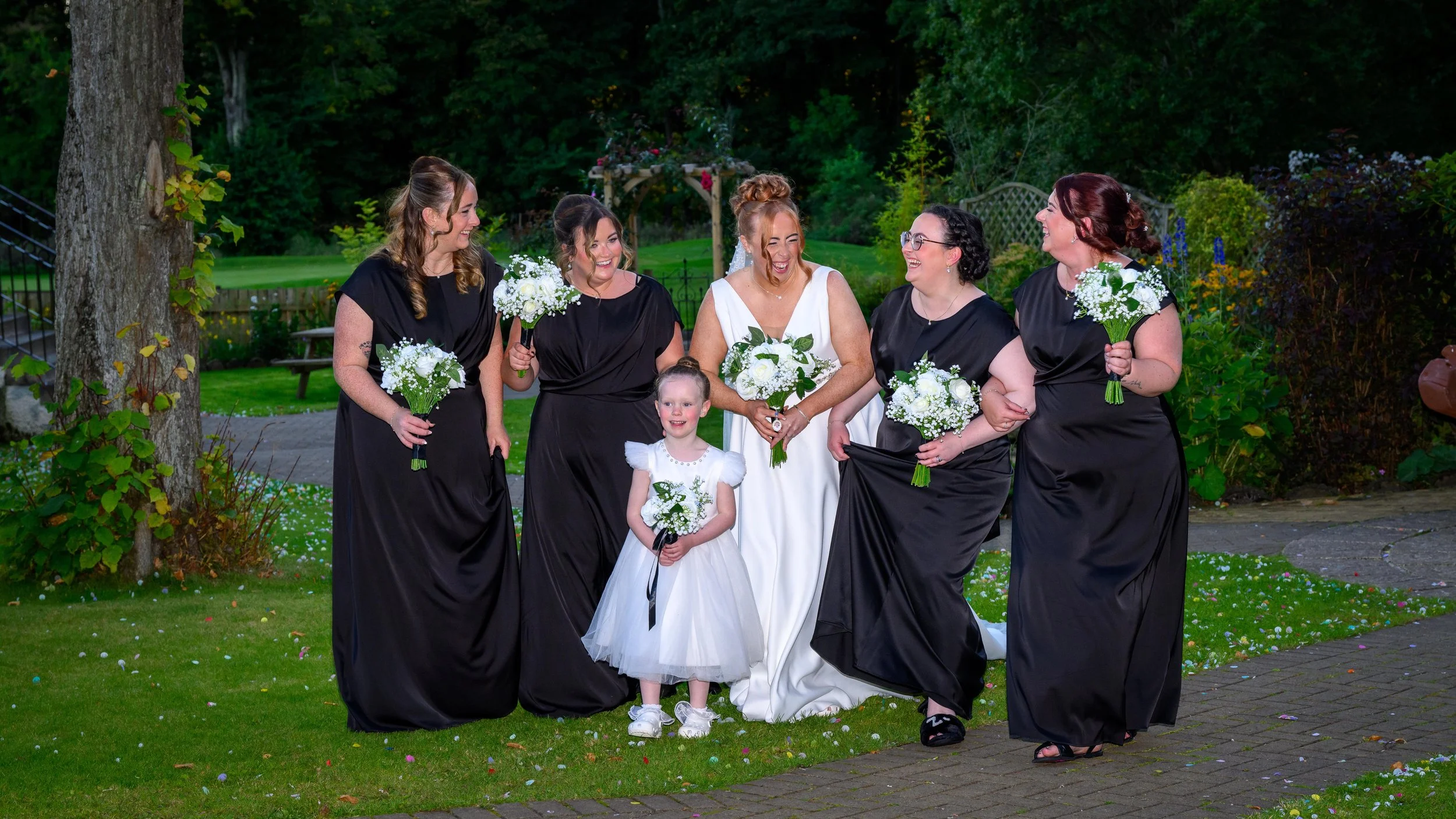 Group of women and a young girl dressed in wedding attire, standing outdoors on a grassy area with trees and flowers in the background. The women are holding bouquets of white flowers, and everyone is smiling and enjoying the moment.