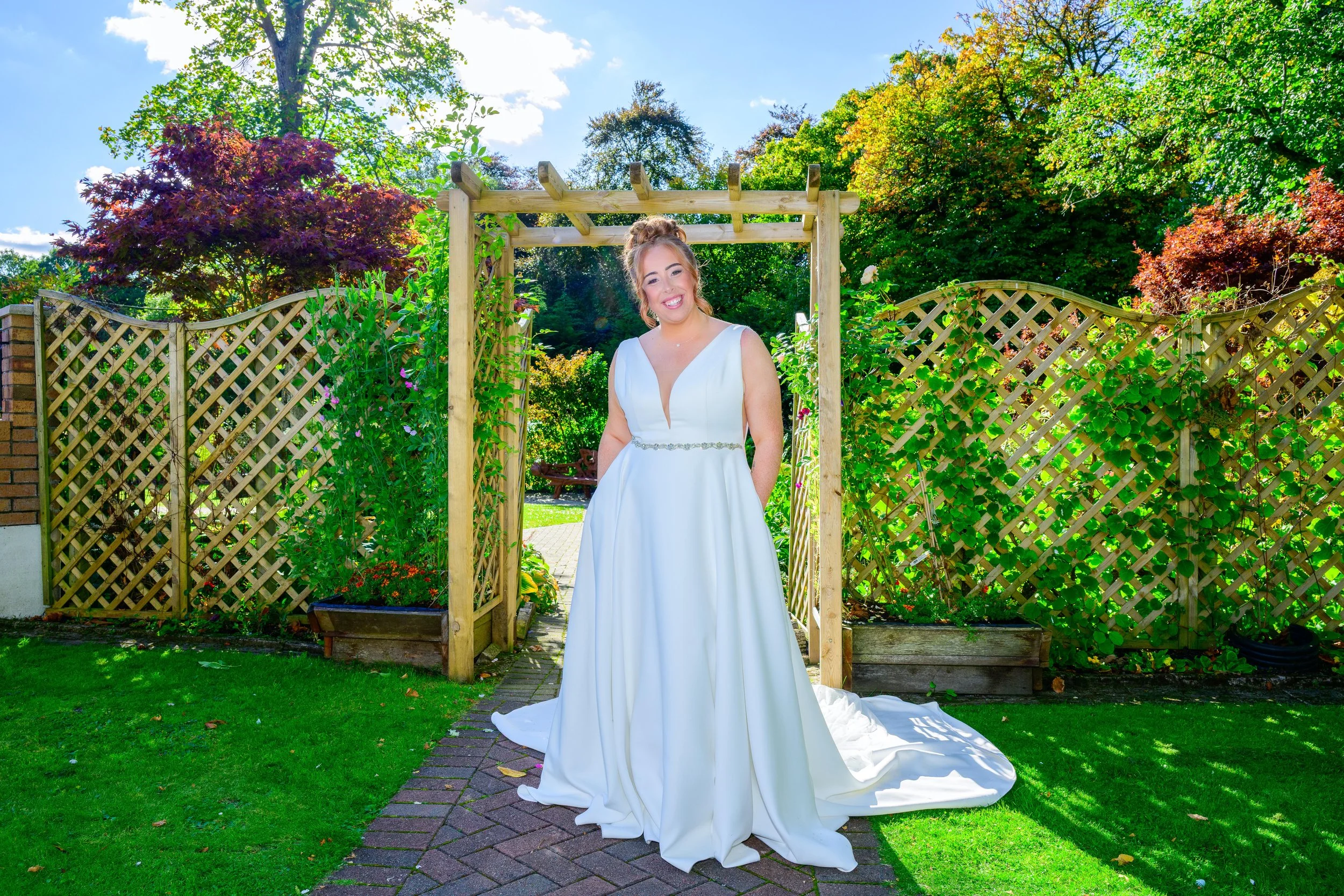 A woman in a white wedding dress standing outdoors in a garden with lush green trees and bushes, a wooden trellis above her, and bright blue sky with scattered clouds in the background.