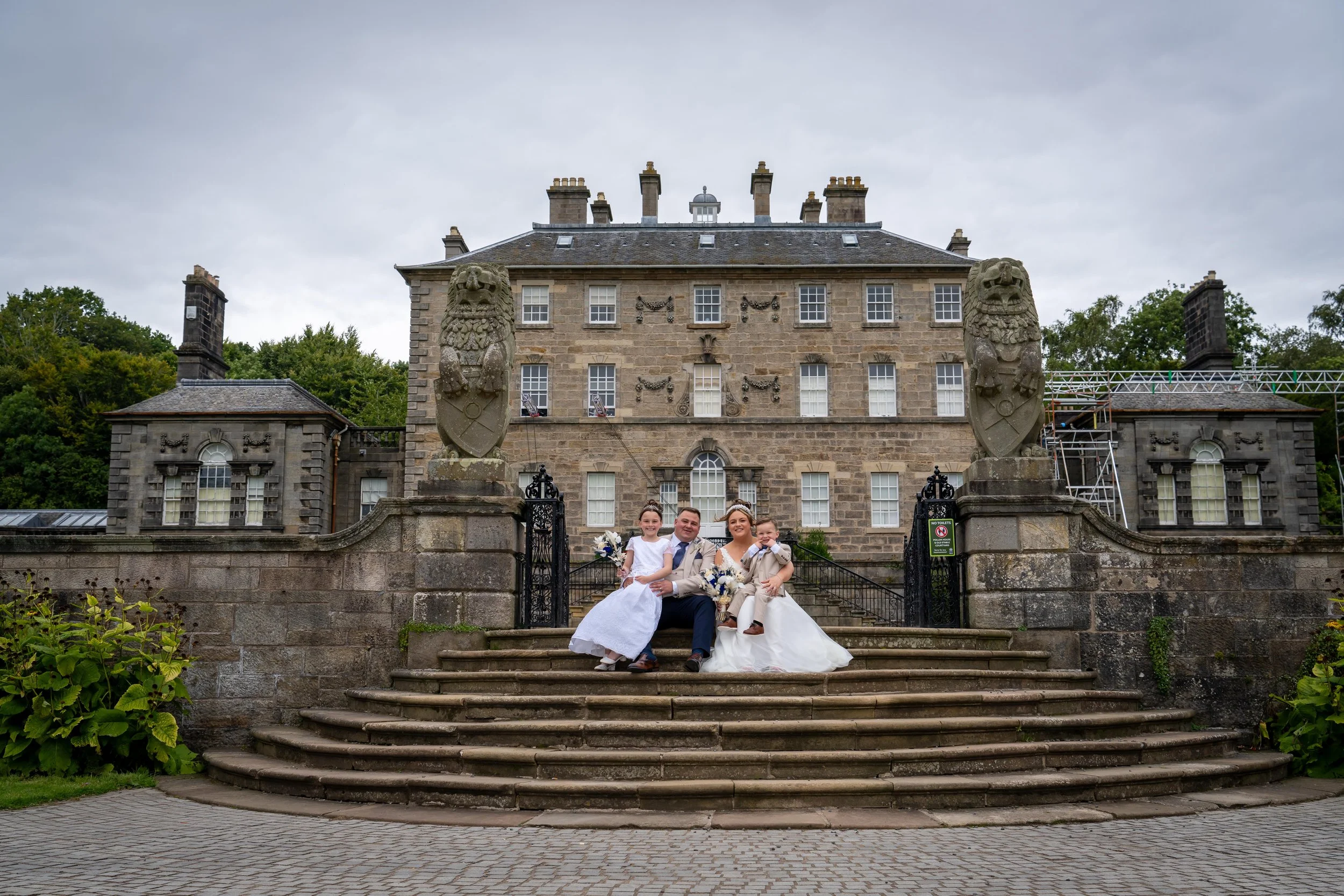 A wedding party with a bride, groom, and two children sitting on stone steps in front of a historic mansion with a stone facade and lion statues at the entrance.