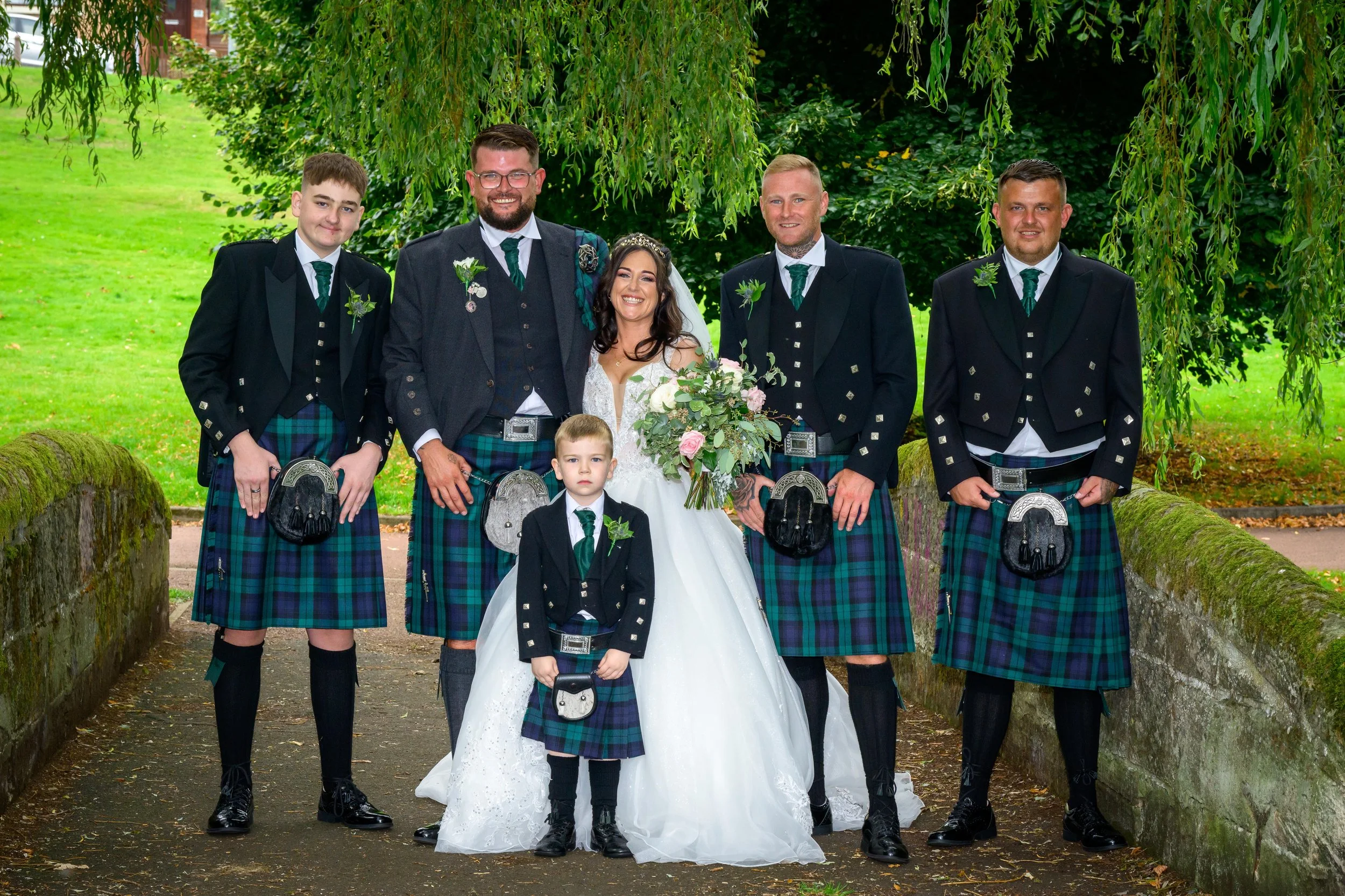 Group of people in traditional Scottish attire, including kilts, posing outdoors on a stone pathway under a tree