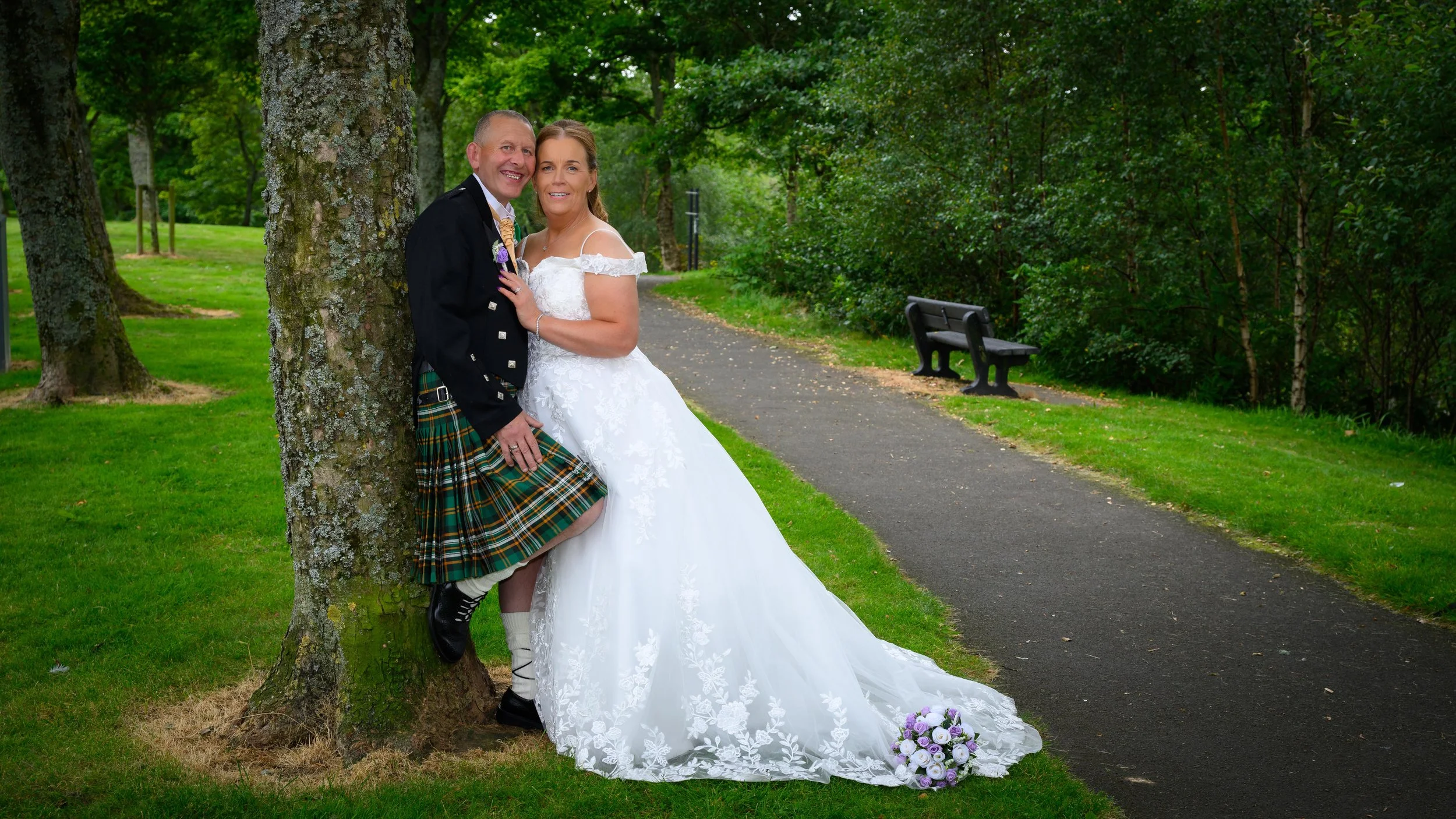 A bride and groom in wedding attire standing next to a tree on a park path, with a bouquet of flowers on the ground nearby.