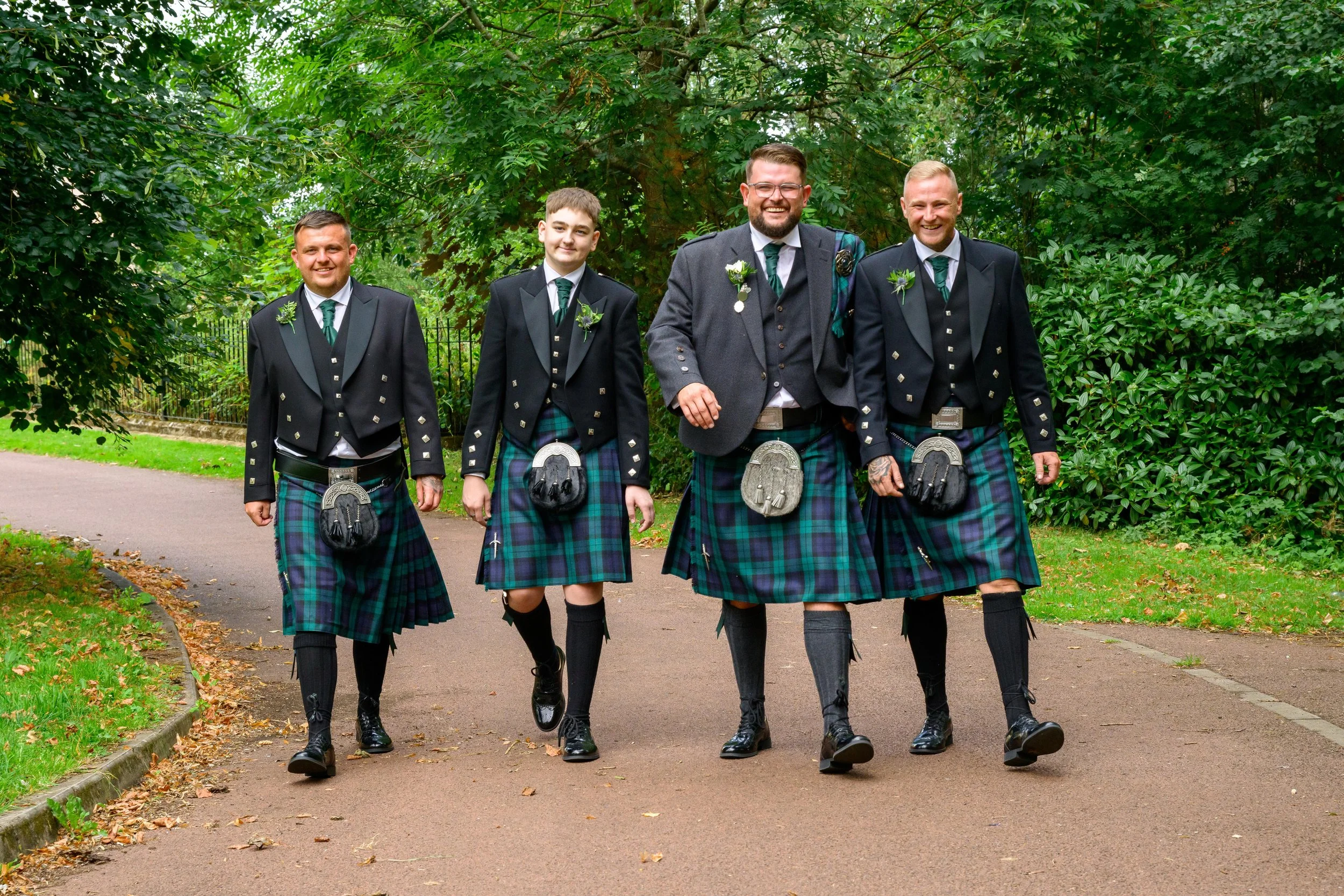 Five men walking outdoors in a park, dressed in traditional Scottish kilts and jackets, smiling.