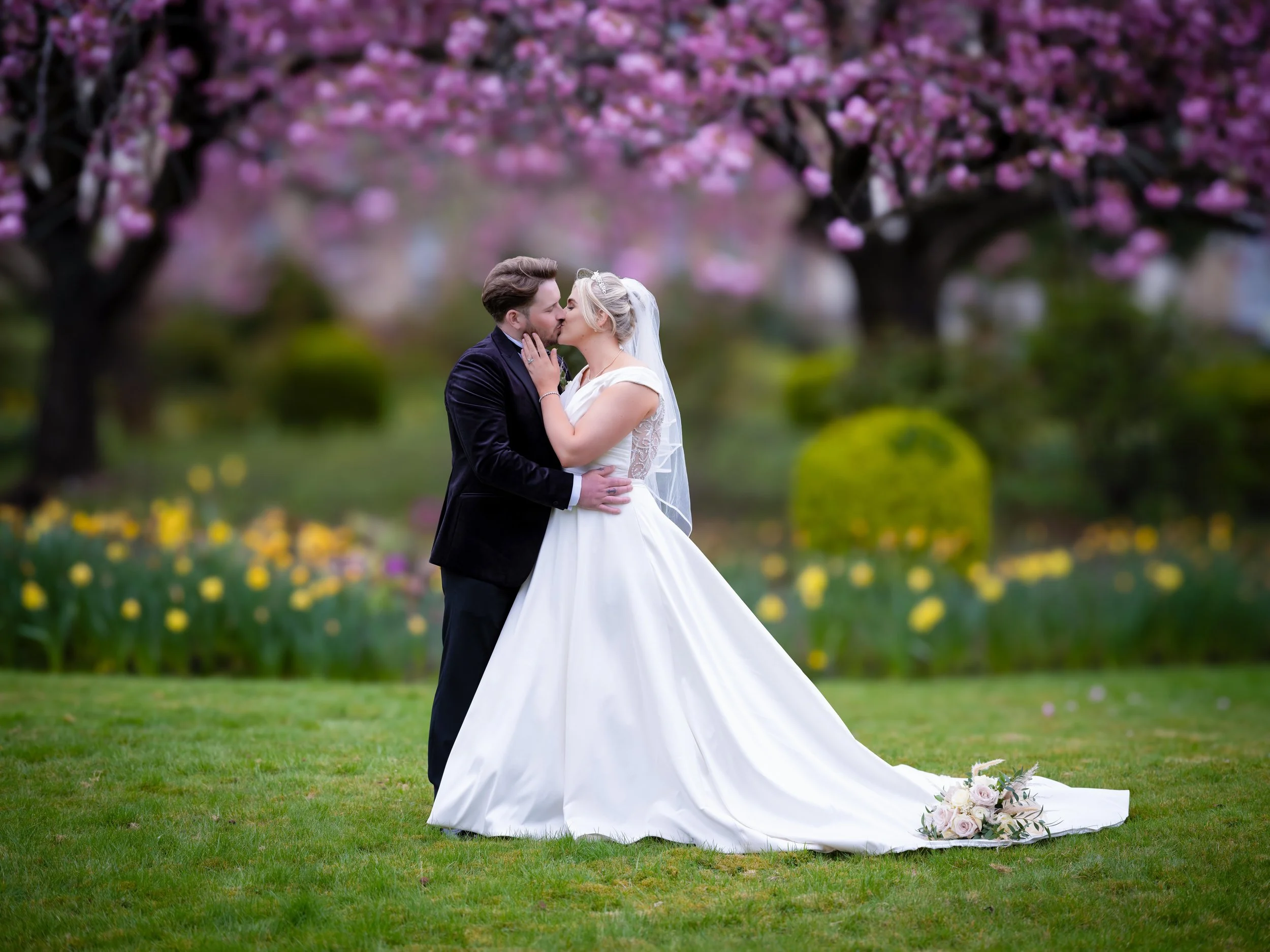 A bride and groom kissing outdoors on a grassy area with pink blossoms in the background, a bouquet of flowers on the ground.